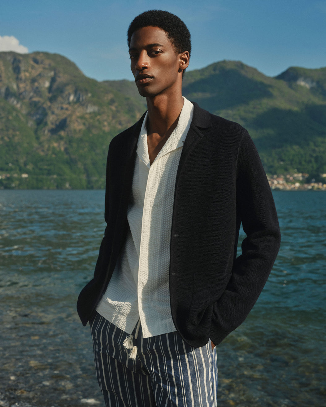 A man stands outdoors by a lake with mountains in the background, wearing a black blazer over a white textured shirt and blue-and-white striped trousers.