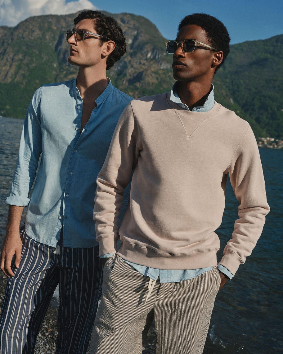 Two men standing outdoors by a lake with mountains in the background, wearing sunglasses, a light blue button-up shirt, and beige and light blue knit tops and striped trousers.