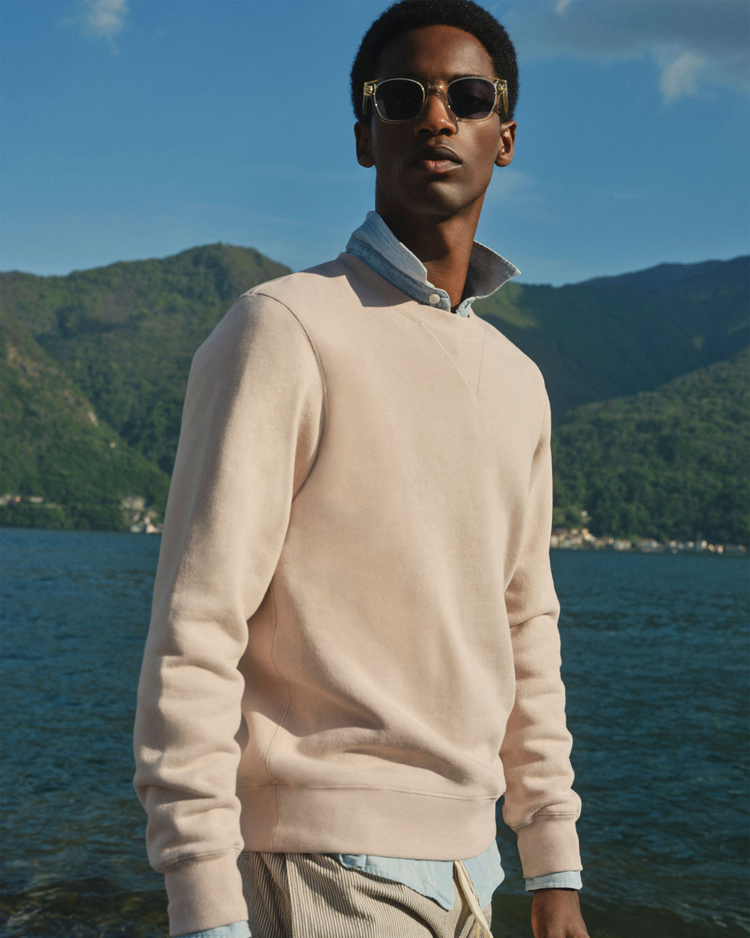 A man wearing a light beige crewneck sweater over a light blue collared shirt, standing outdoors by a lake with mountains in the background.