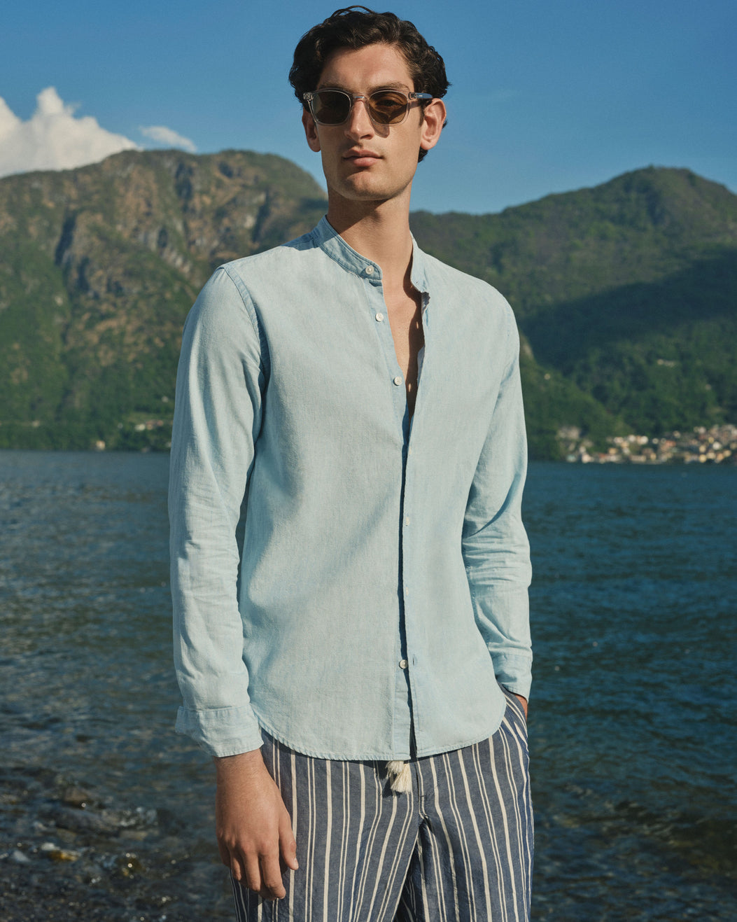 A man wearing a light blue long-sleeve linen shirt and striped trousers stands by a lake with mountains in the background.