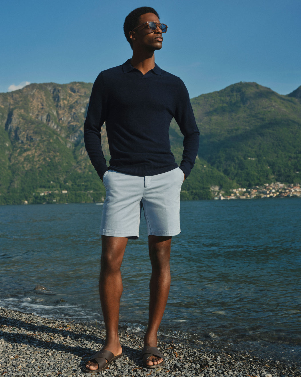 A man wearing a navy long-sleeve polo shirt, light blue shorts, and brown sandals stands on a pebble shore with water and green mountains in the background.