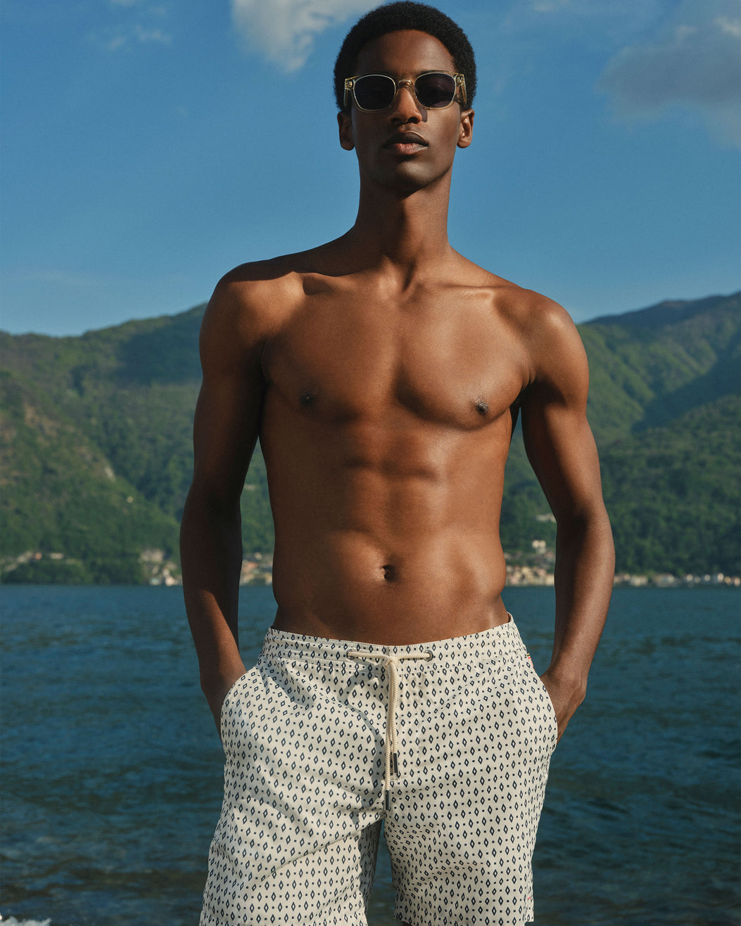 A shirtless man wearing patterned beige swim trunks and sunglasses stands outdoors by the water with mountains in the background.