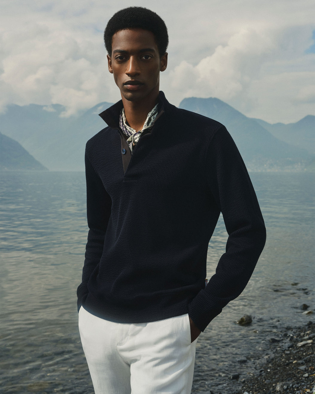 A man standing outdoors by a lake with mountains and cloudy sky in the background, wearing a dark navy quarter-zip sweater over a patterned collared shirt and white trousers.