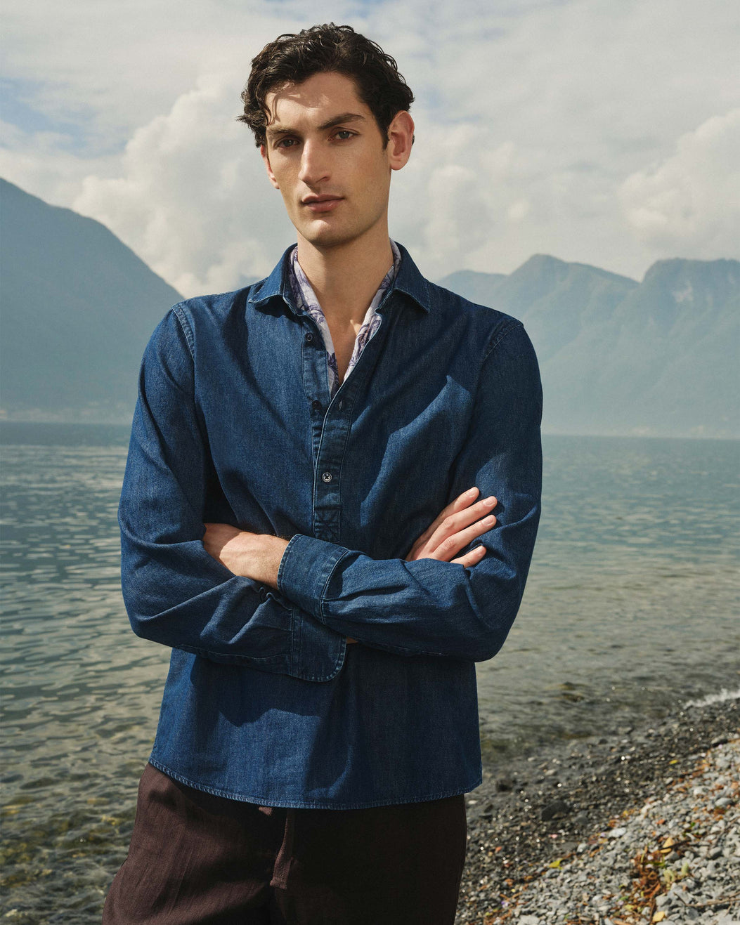 Man standing with arms crossed by a body of water with mountains in the background
