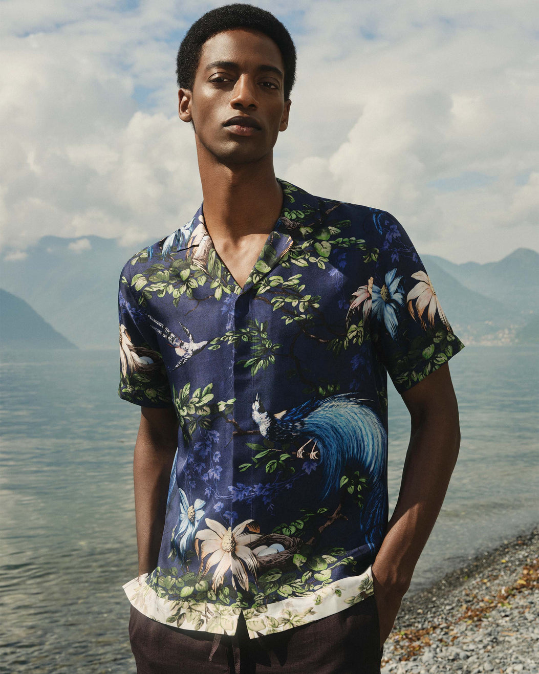 Man wearing a floral shirt by a lakeside with mountains in the background