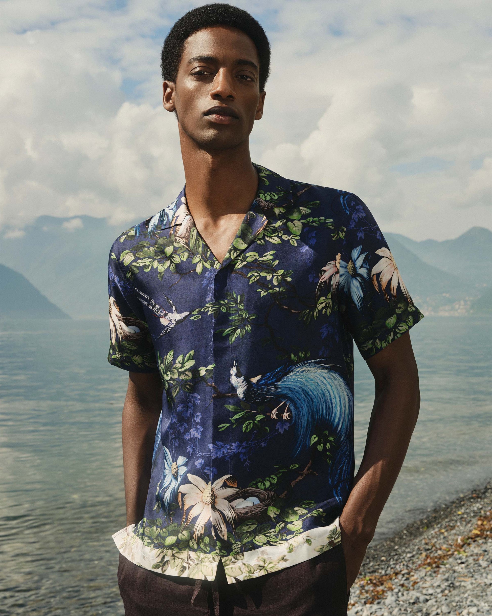 Man wearing a floral shirt by a lakeside with mountains in the background