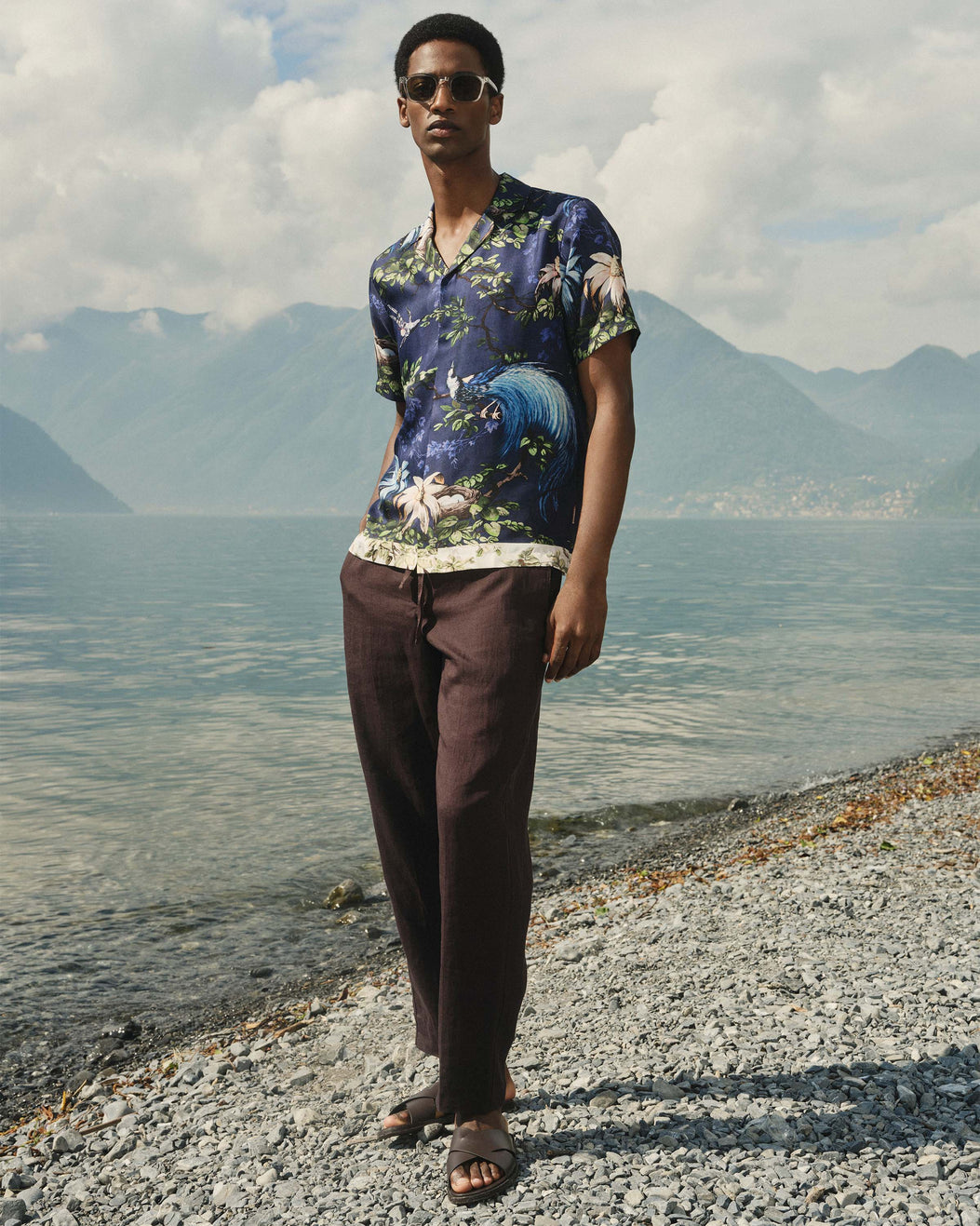 Man wearing a patterned shirt and sunglasses standing by a lake with mountains in the background