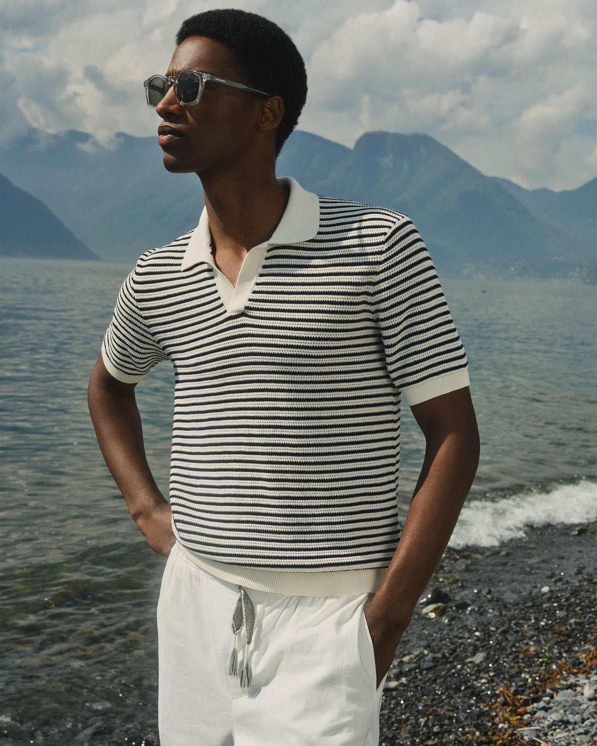 A man wearing a black and white horizontally striped short-sleeve knit polo shirt and white drawstring shorts, standing on a pebble beach with mountains and water in the background.