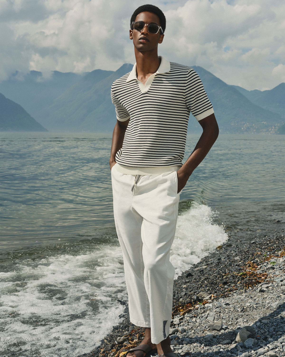 A man wearing a black and white striped short-sleeve polo shirt and white drawstring trousers stands on a pebble shore with water and mountains in the background.