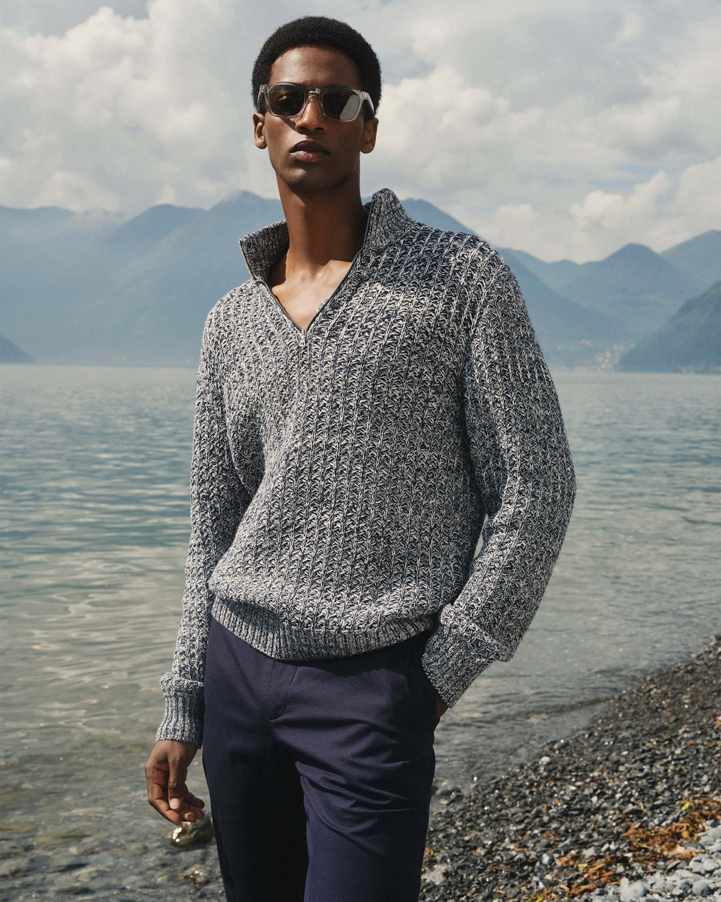 Man wearing a gray sweater and sunglasses standing by a lake with mountains in the background