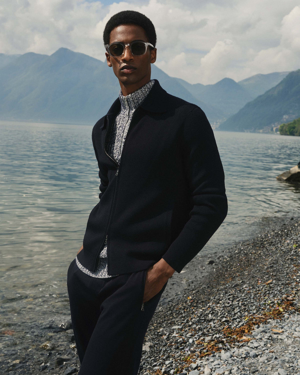 Man in a black jacket and sunglasses standing by a lake with mountains in the background