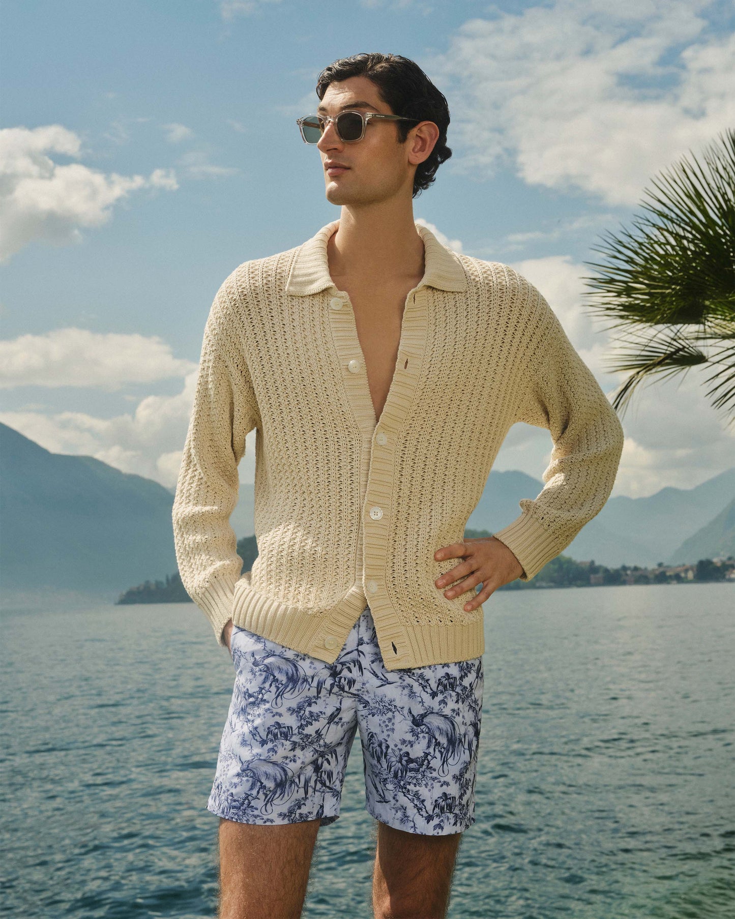 Man wearing a beige cardigan and patterned shorts standing by a lake with mountains in the background.