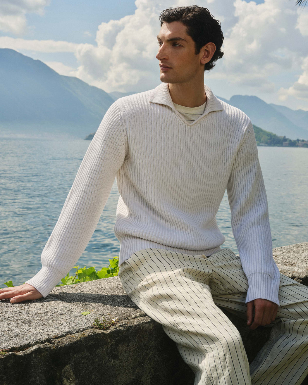 Man sitting by a lake with mountains in the background, wearing a white sweater and light-colored pants.