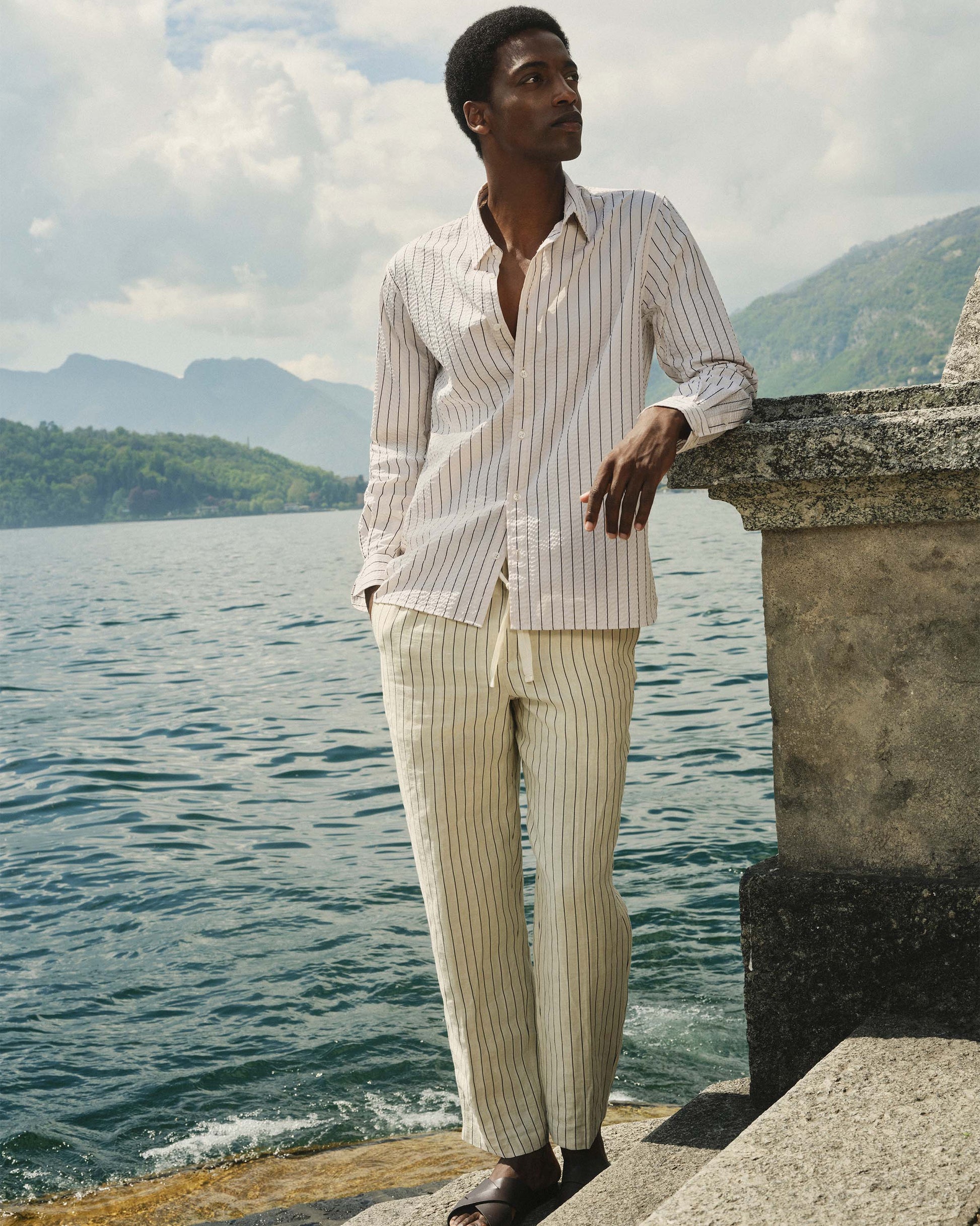 Man in striped shirt and pants standing by a body of water with mountains in the background