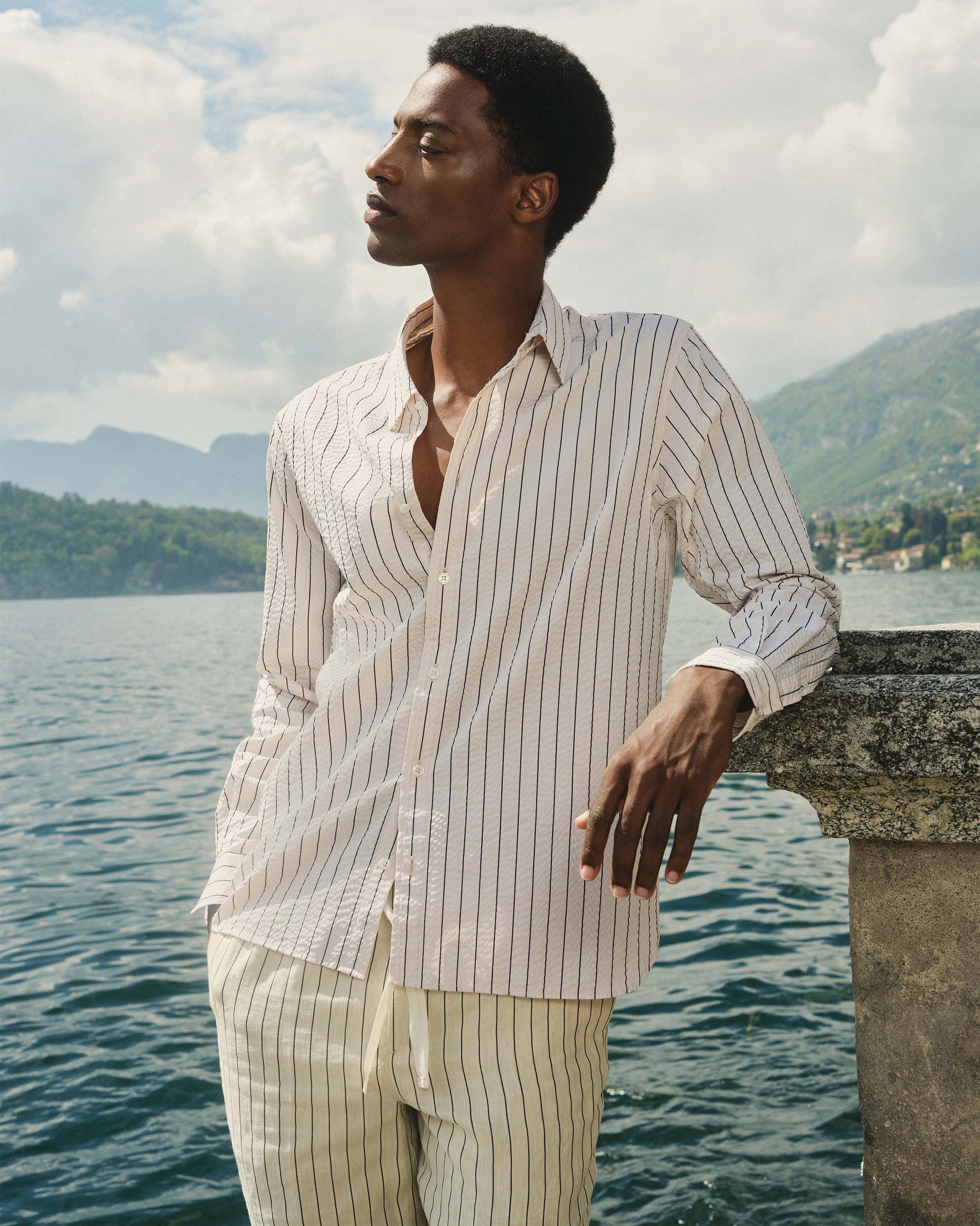 Man in a striped shirt and pants standing by a body of water with mountains in the background