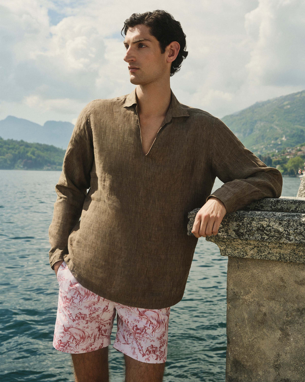 Man wearing a brown shirt and pink shorts with floral pattern by a body of water and mountains.