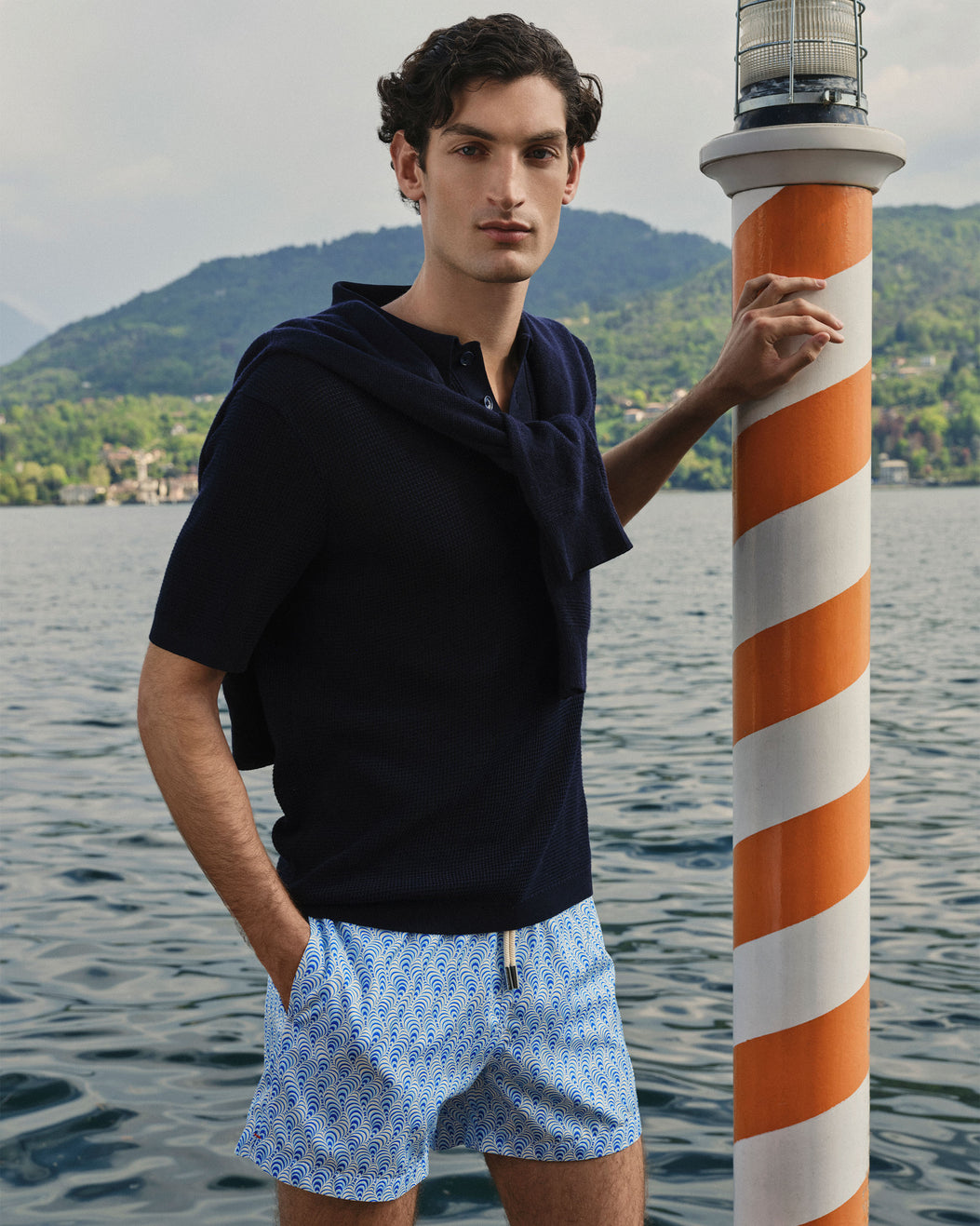 A man stands by a lake wearing a dark navy short-sleeve polo shirt and light blue patterned swim shorts, with a large orange-and-white striped pole beside him and mountains in the background.