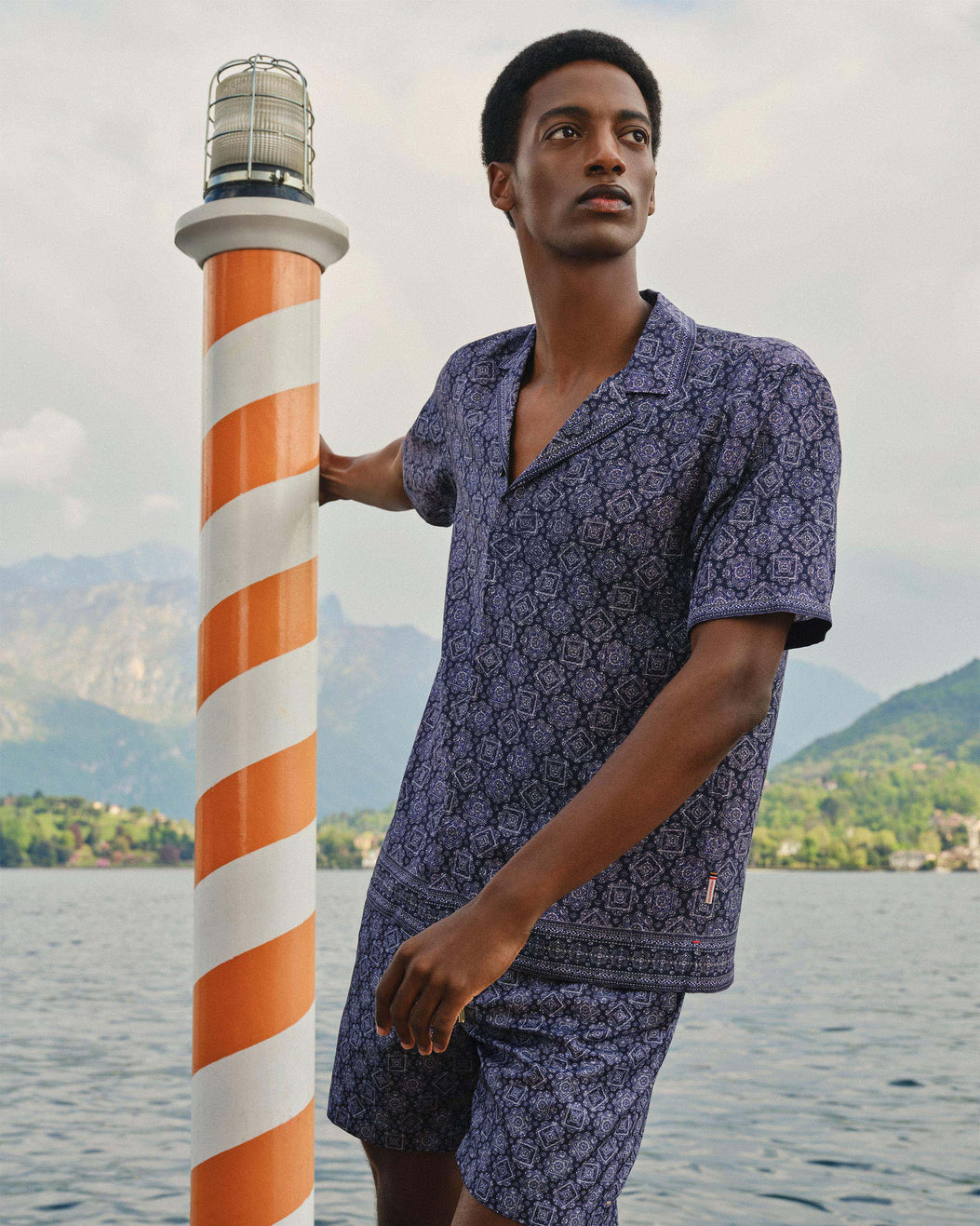 A man wearing a matching short-sleeve button-up shirt and shorts set in a blue and white floral pattern, standing next to a striped orange and white lighthouse post with a lake and mountains in the background.