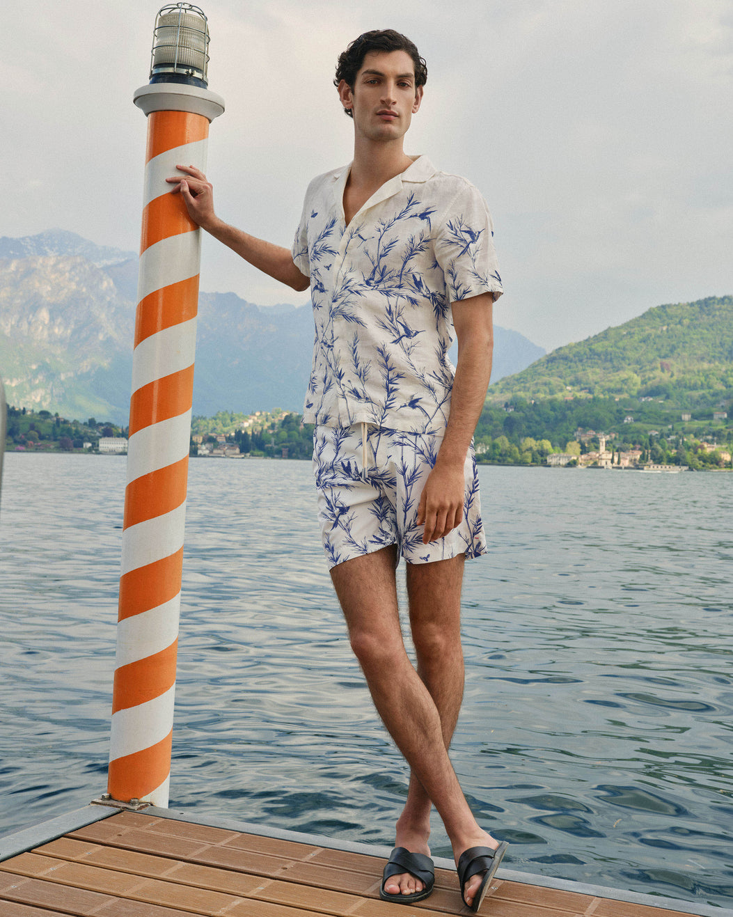 A man stands on a wooden dock by a lake wearing a white short-sleeve shirt and matching shorts with a blue botanical print. He is posing next to a striped orange and white pole with a lighthouse fixture, with mountains and water in the background.