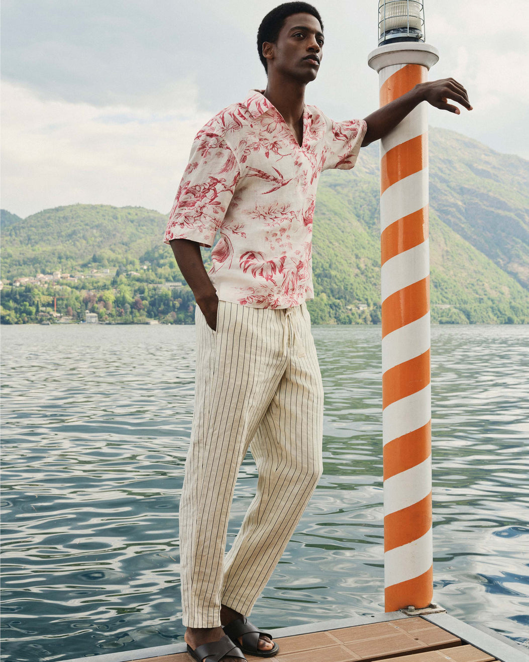 Man in a floral shirt and striped pants standing by a striped pole on a dock with mountains in the background.