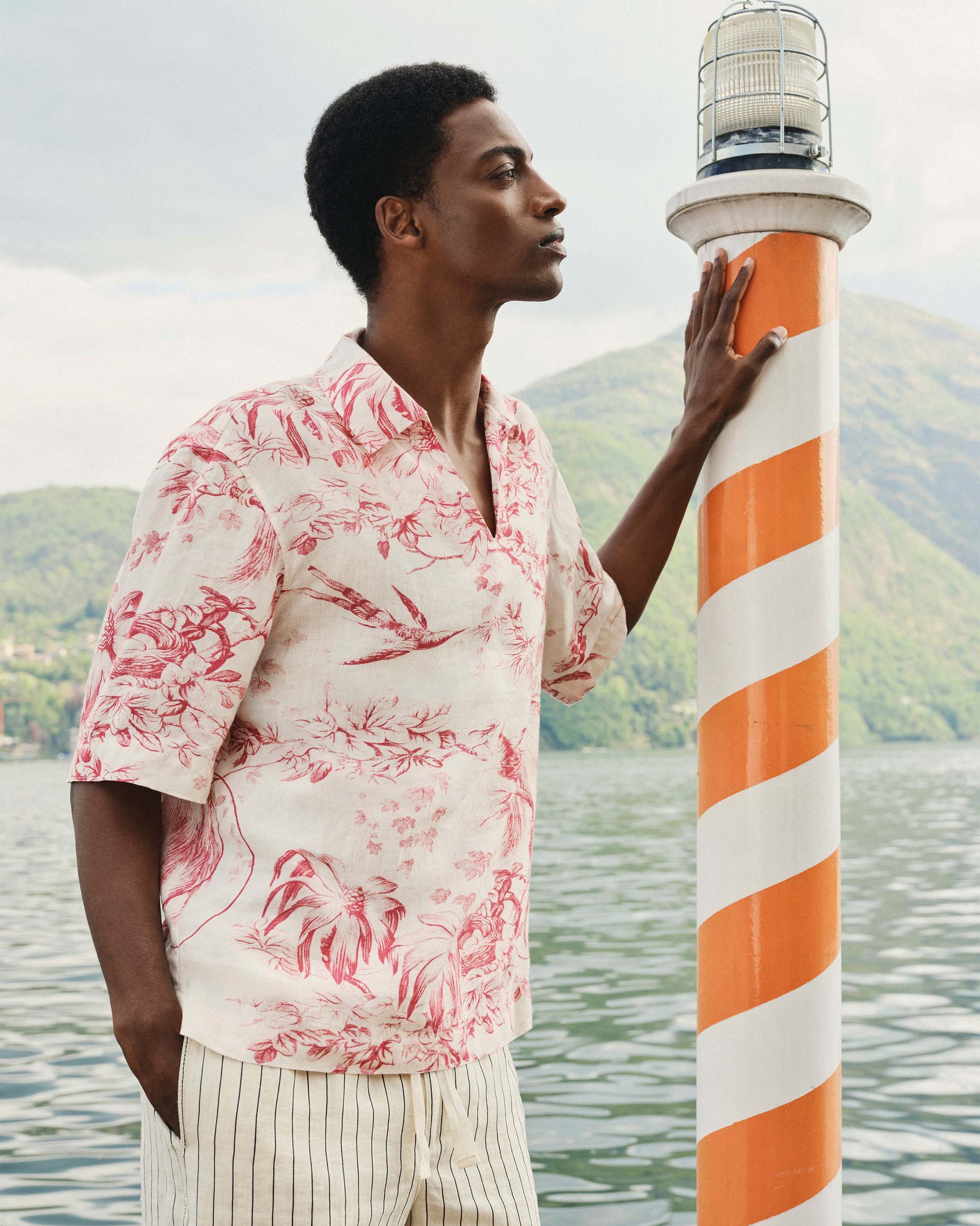 Man in a floral shirt standing next to a striped lighthouse by a body of water.