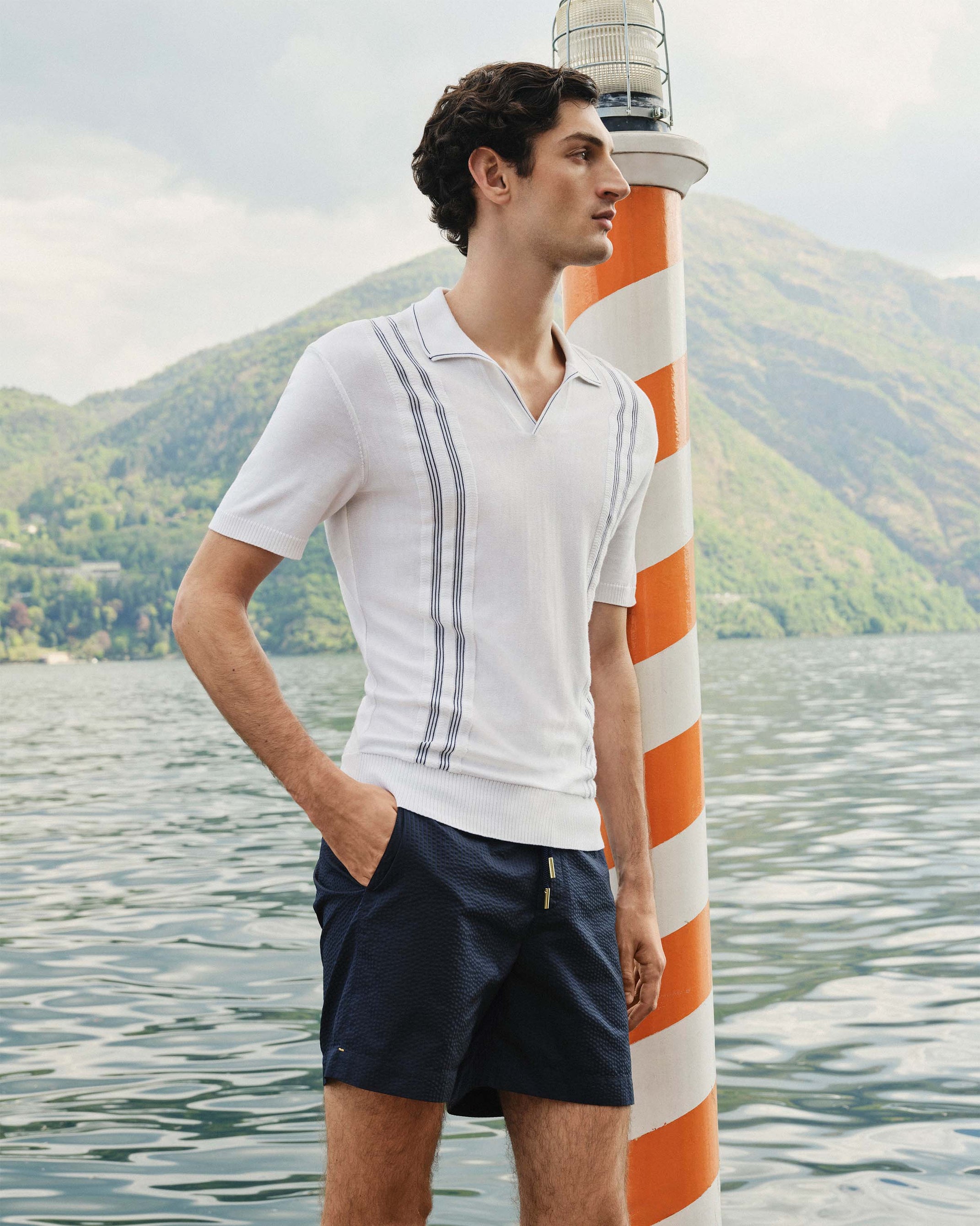 Man in a white polo shirt and navy shorts standing by a striped lighthouse by a lake.