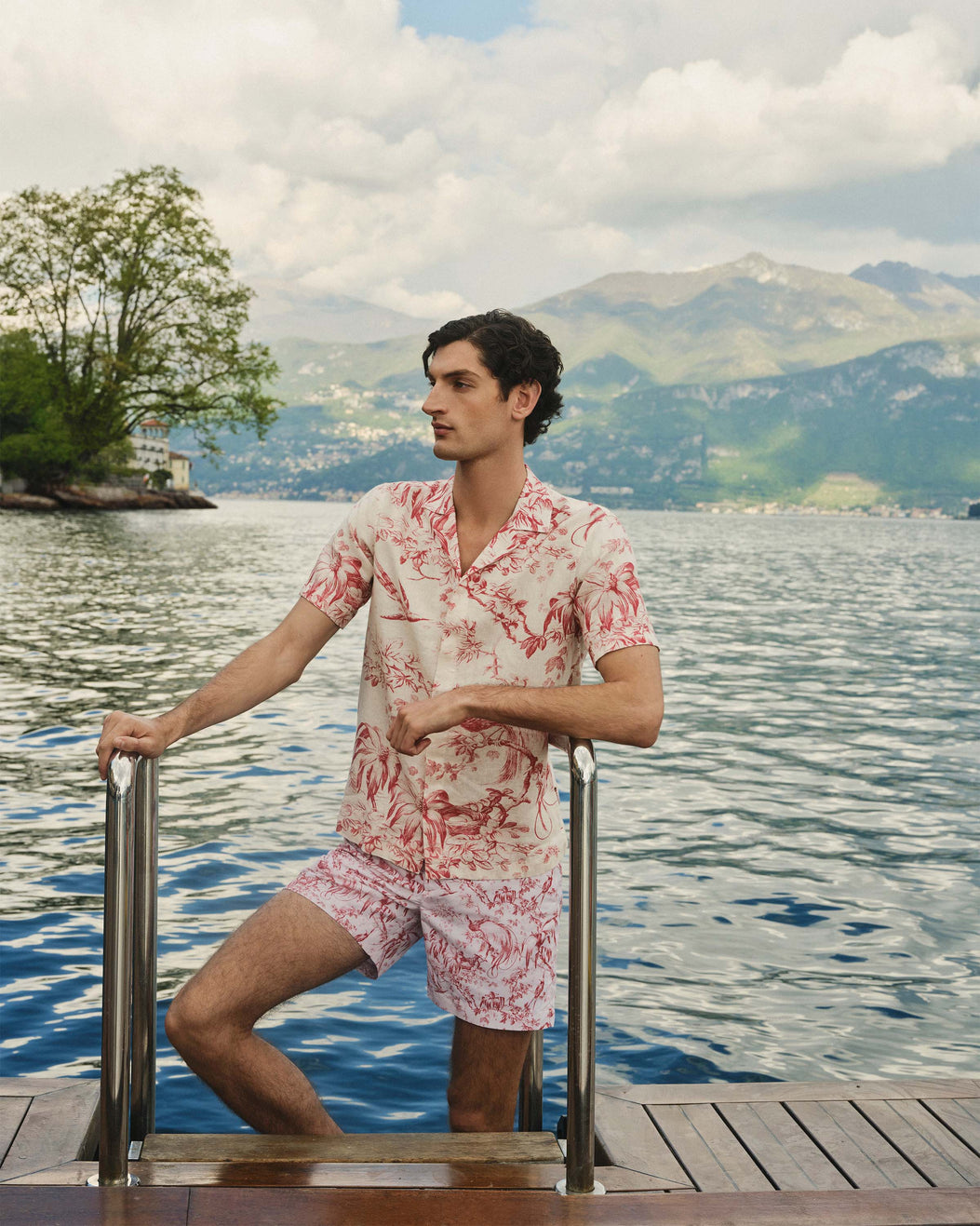 Man in a floral outfit standing on a dock by a lake with mountains in the background