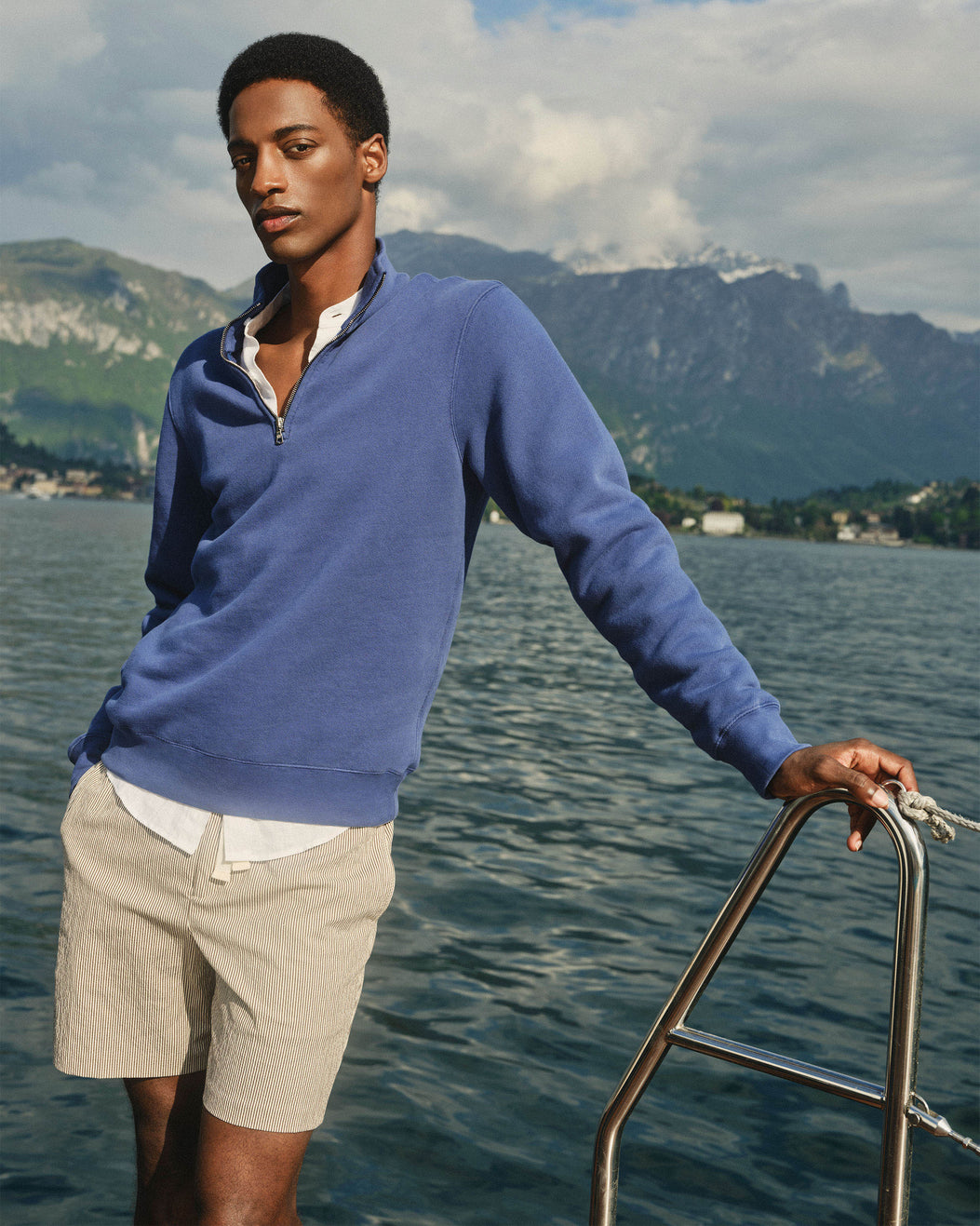 A man wearing a blue quarter-zip long-sleeve sweater and beige shorts stands on a boat with water and mountains in the background.