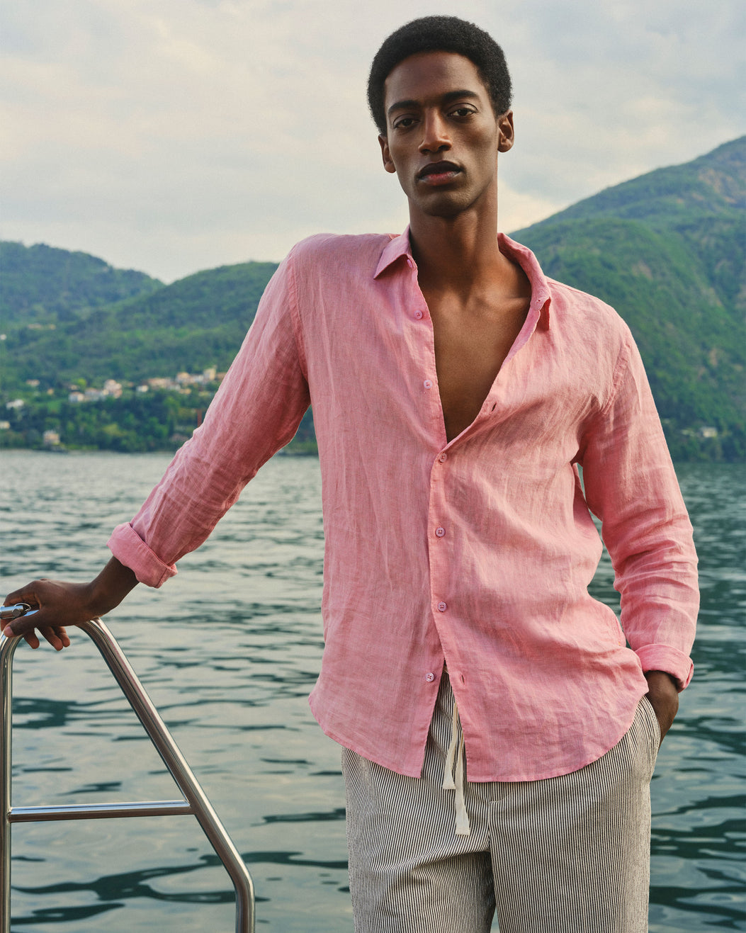 A man wearing a light pink linen button-up shirt and beige trousers stands on a boat with water and green hills in the background.
