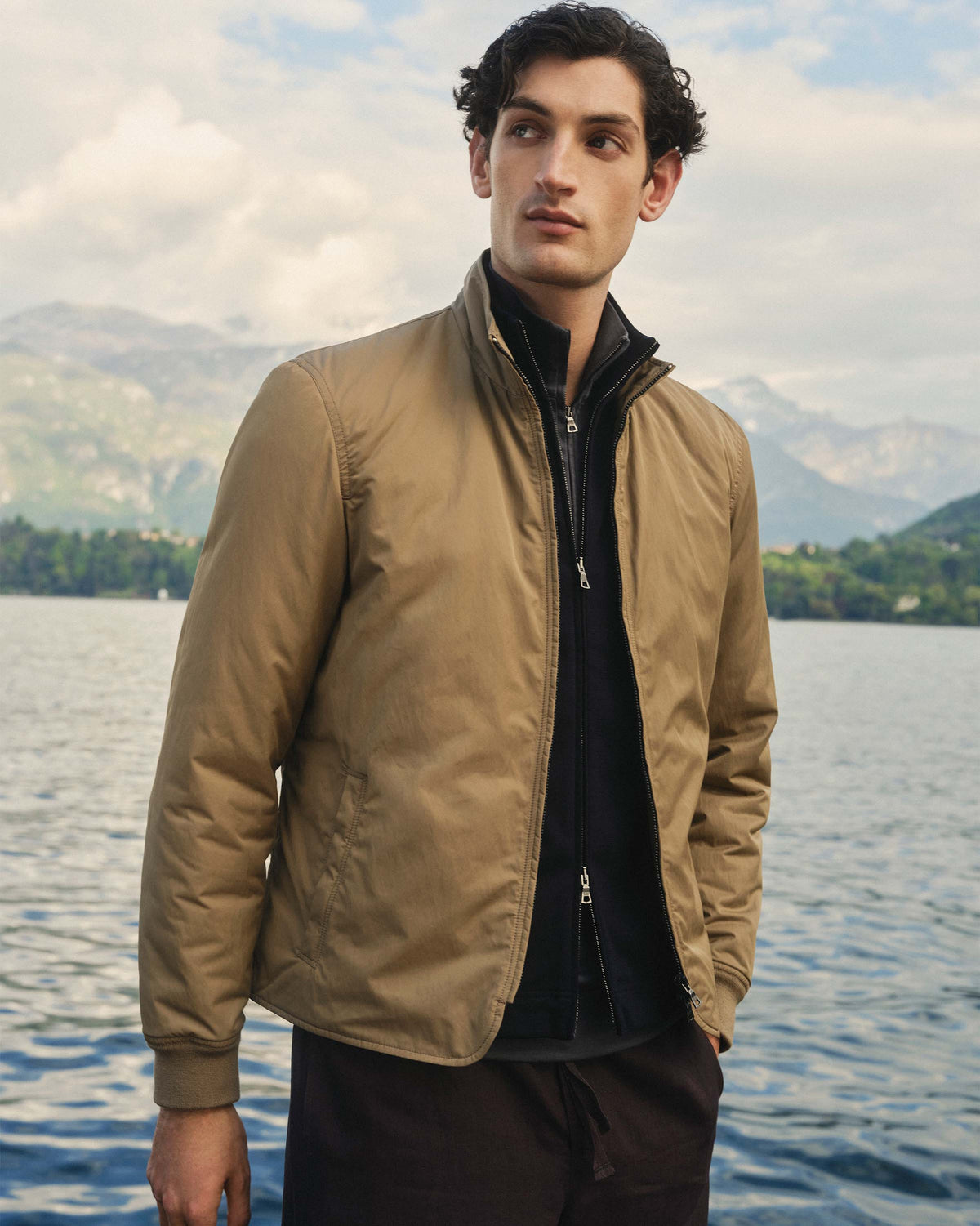Man wearing a beige jacket standing by a lake with mountains in the background
