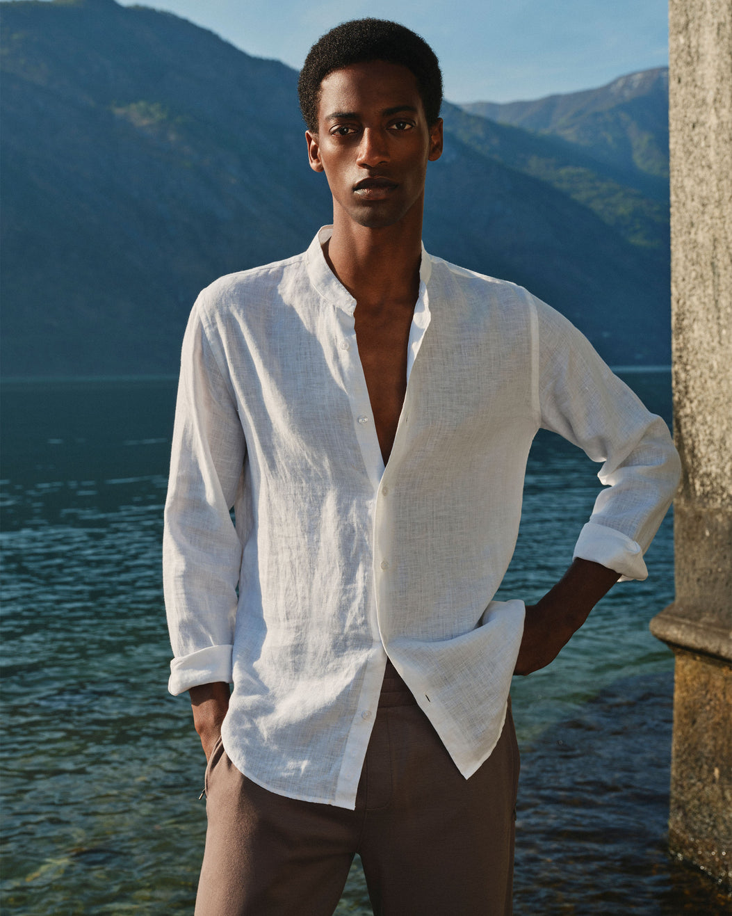 A man wearing a white linen button-down shirt and brown trousers stands outdoors by a lake with mountains in the background.