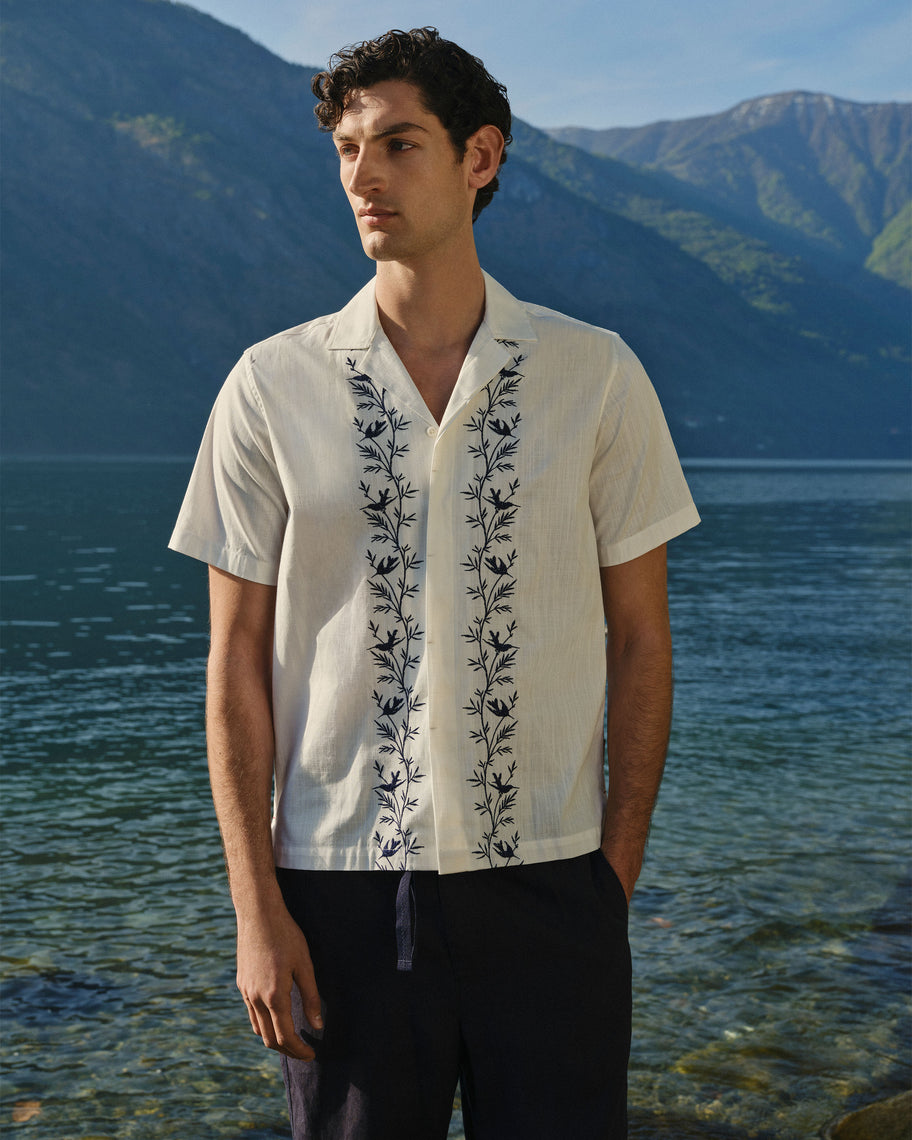 A man wearing a short-sleeve cream-colored button-up shirt with dark blue floral embroidery down the front paired with black trousers standing outdoors in front of a scenic mountain and lake background.