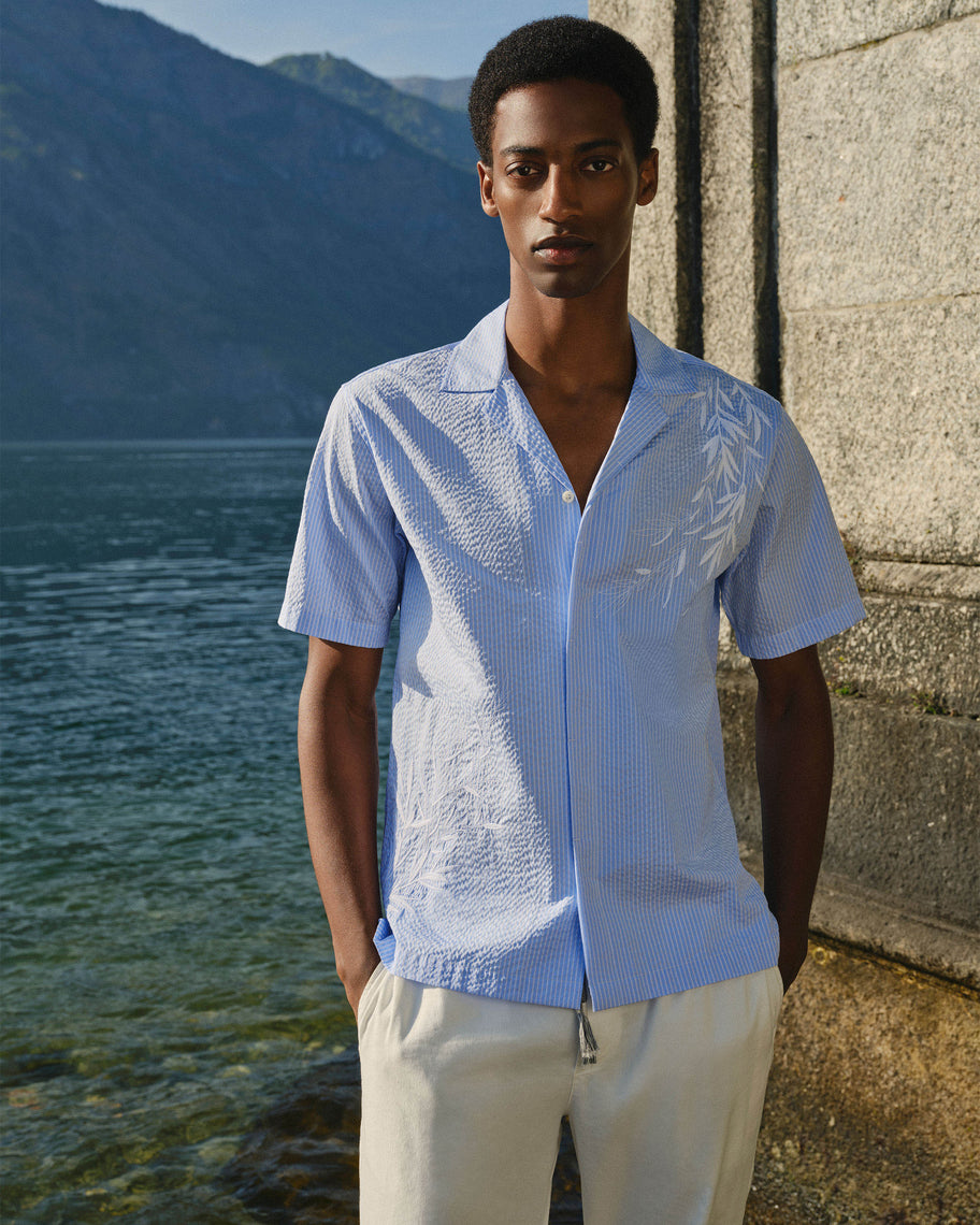 A man stands outdoors beside a stone wall and water, wearing a light blue short-sleeve button-front shirt with a subtle textured pattern and white pants.