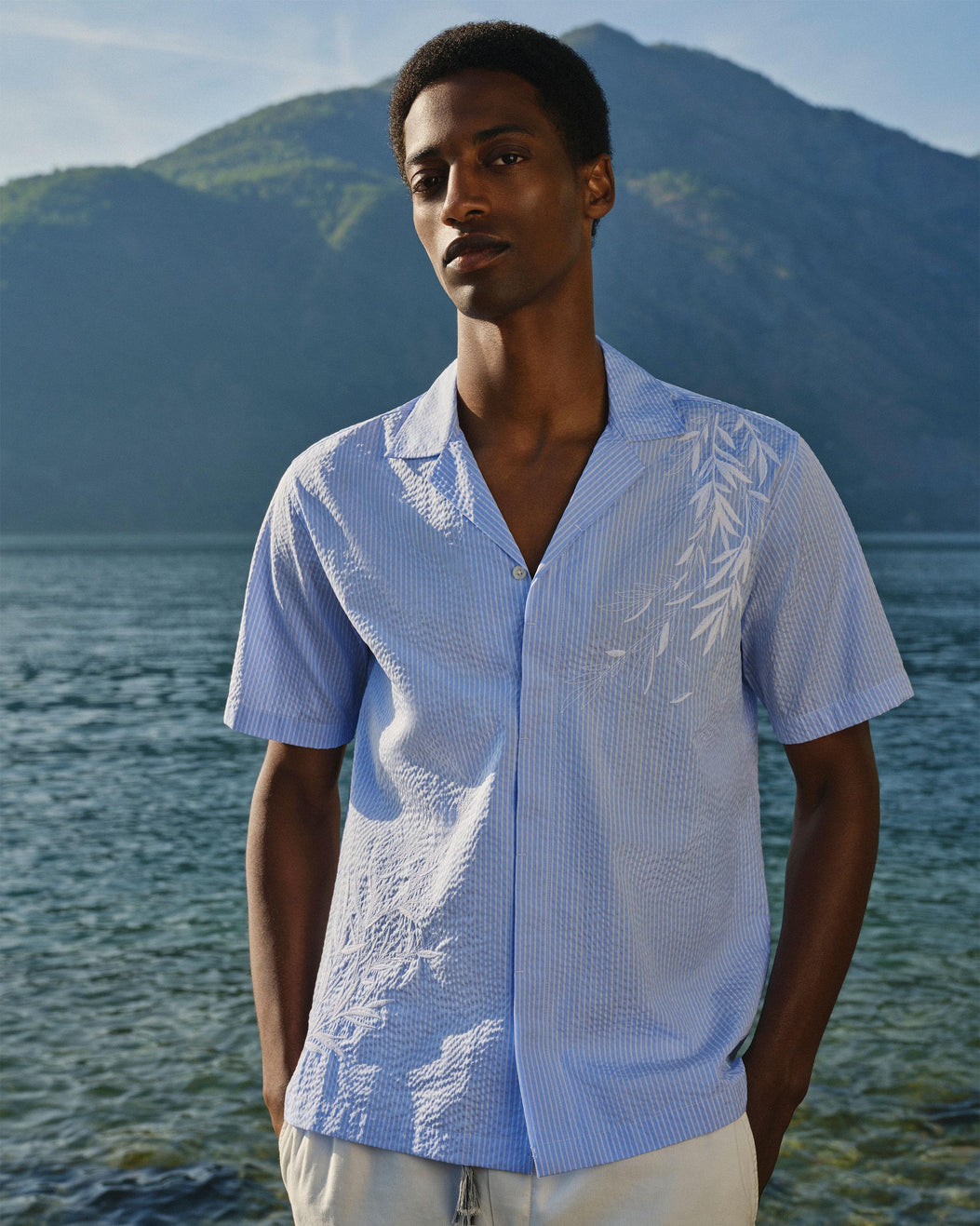 A man wearing a light blue short-sleeve button-up shirt with white leaf embroidery paired with white trousers,  standing outdoors in front of a lake and green mountains.