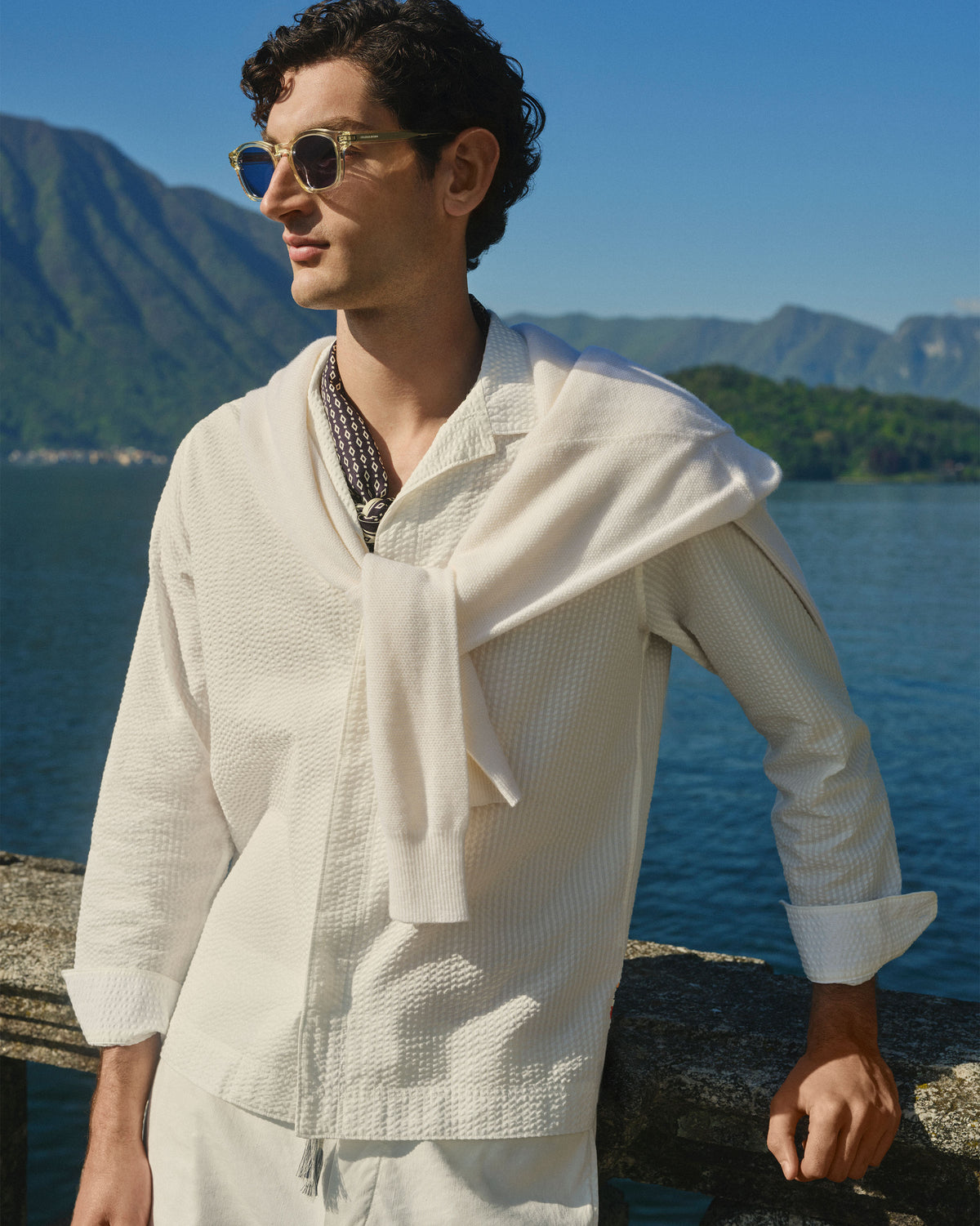 A man wearing a textured off-white long-sleeve shirt and matching shorts stands outdoors by a lake with mountains in the background. He is wearing round sunglasses and a patterned necklace.