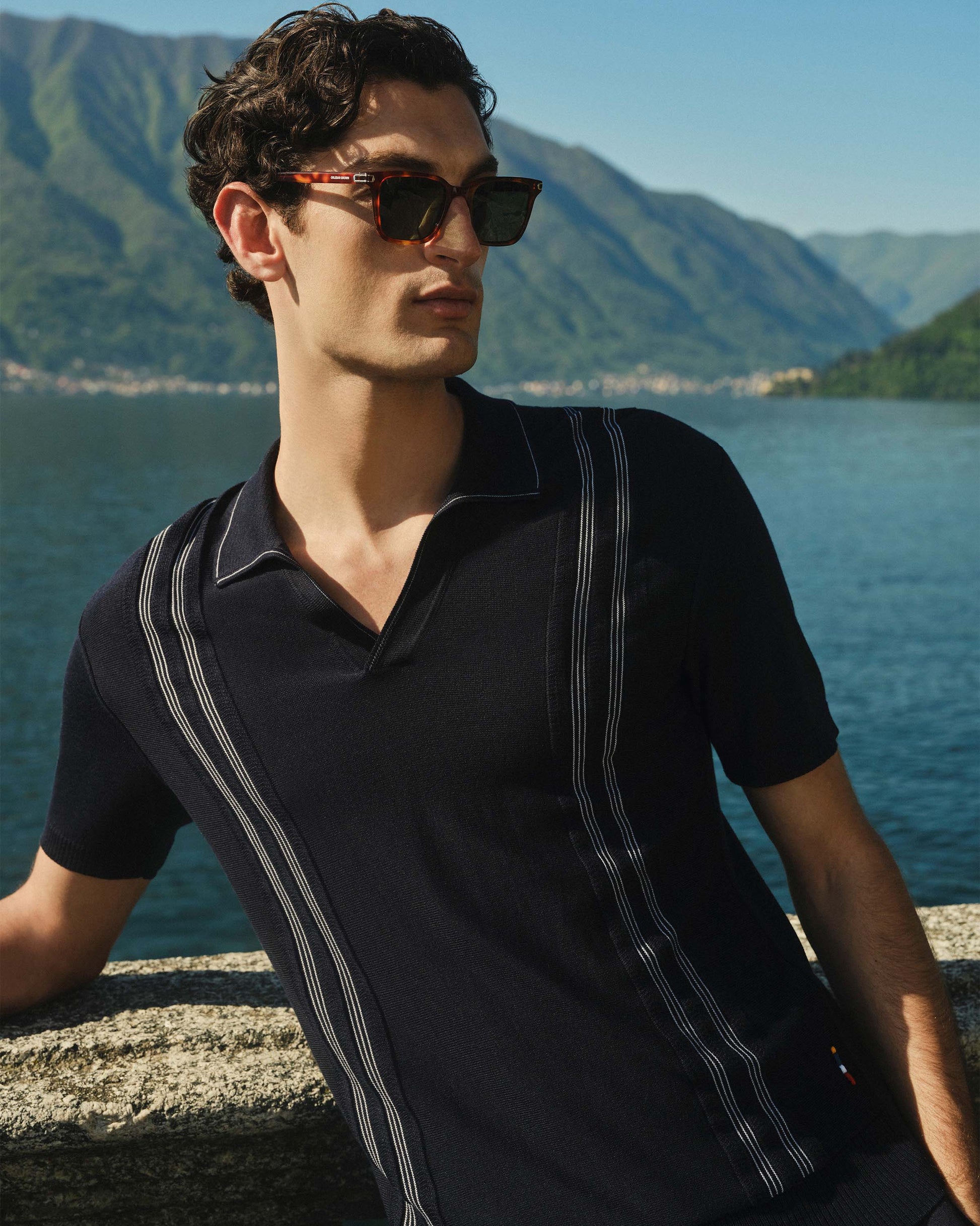 Man wearing sunglasses and a black polo shirt by a scenic lake with mountains.