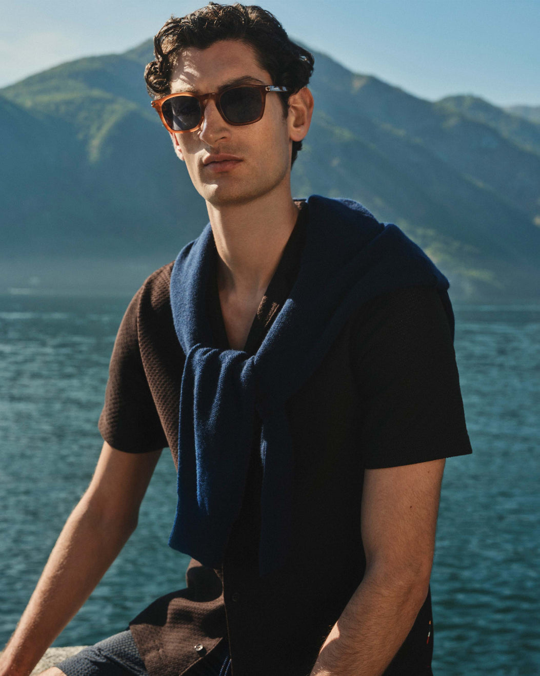 Man wearing sunglasses and a dark shirt with a blue towel draped over his shoulder, standing against a mountain and water backdrop.
