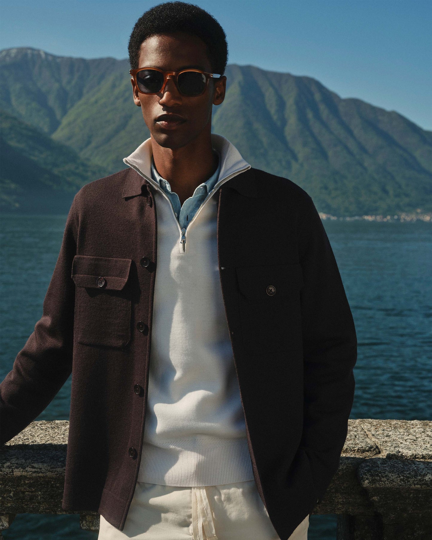 Man wearing sunglasses and a brown jacket with a mountain and lake in the background