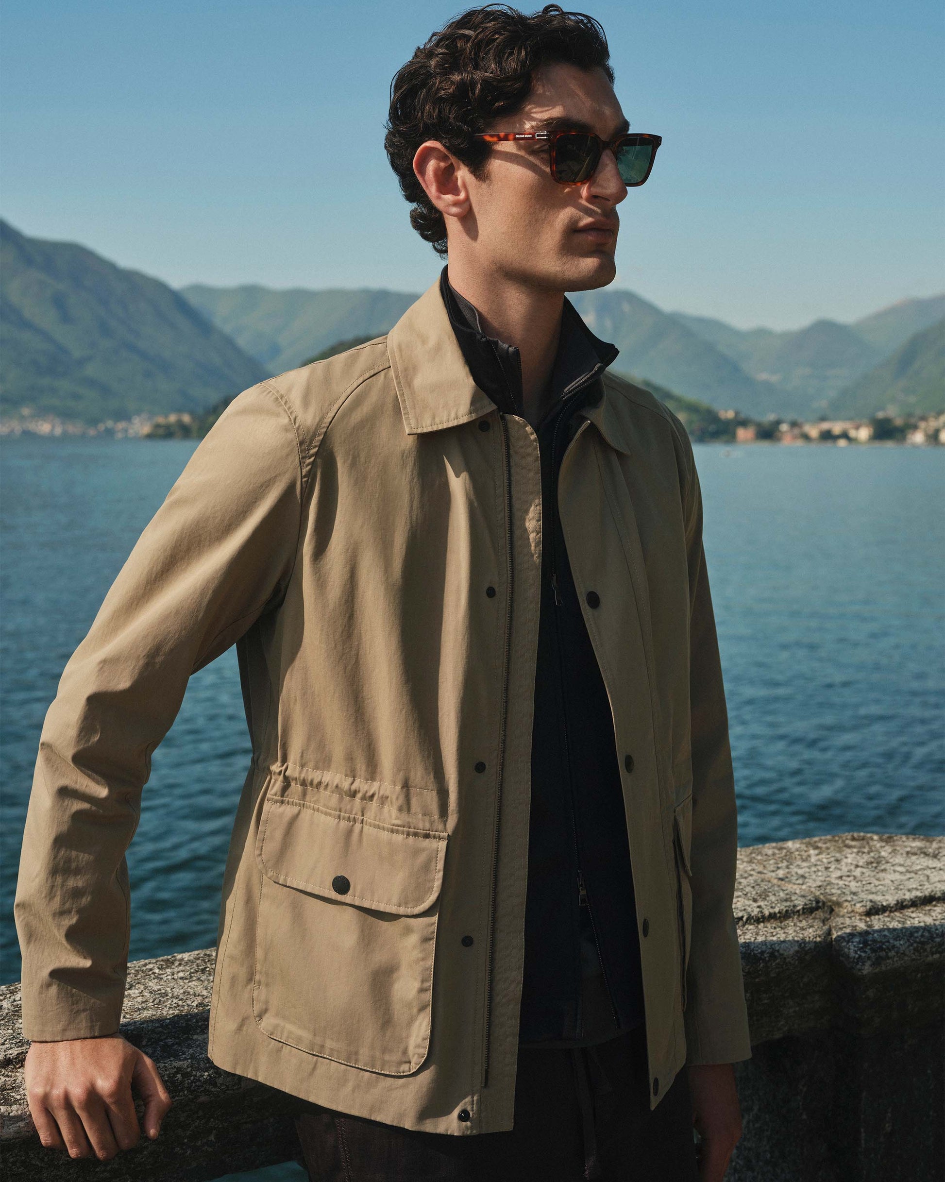 Man wearing a beige jacket and sunglasses by a body of water with mountains in the background
