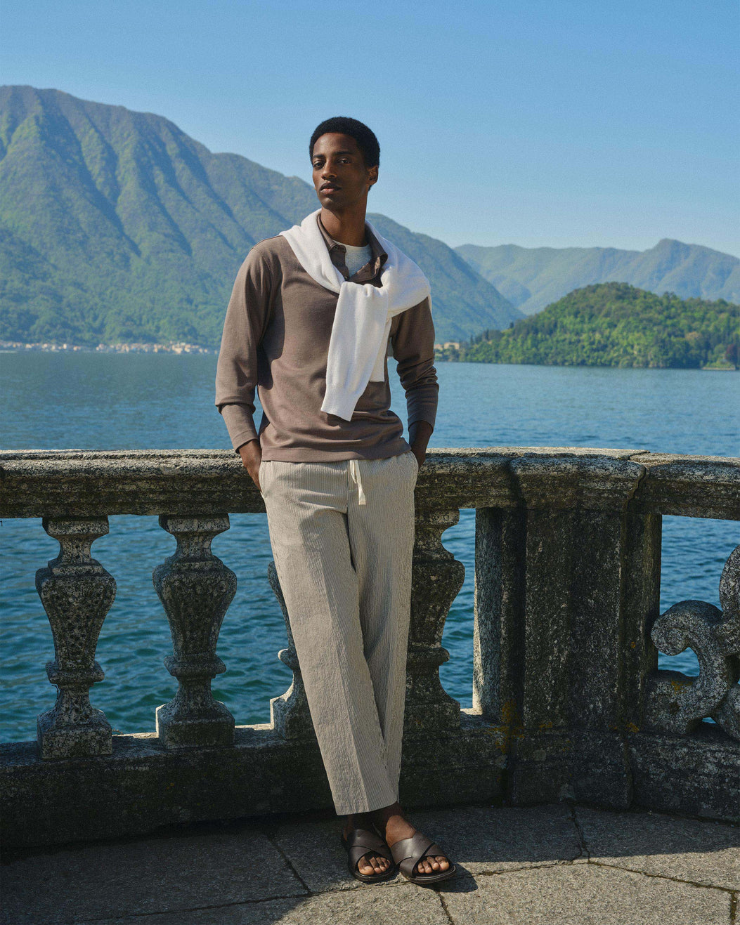 A man stands outdoors by a stone railing overlooking a lake with mountains in the background, wearing a taupe knit sweater, light beige drawstring trousers, a white draped scarf, and dark brown sandals.