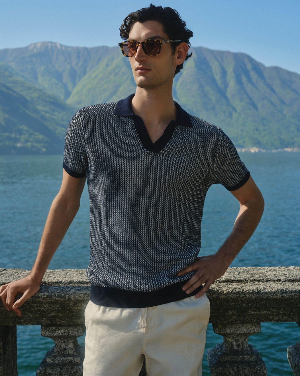A man wearing a navy and beige textured knit polo shirt with a dark collar paired wit h white trousers standing on a stone balcony with a scenic mountain and lake background.