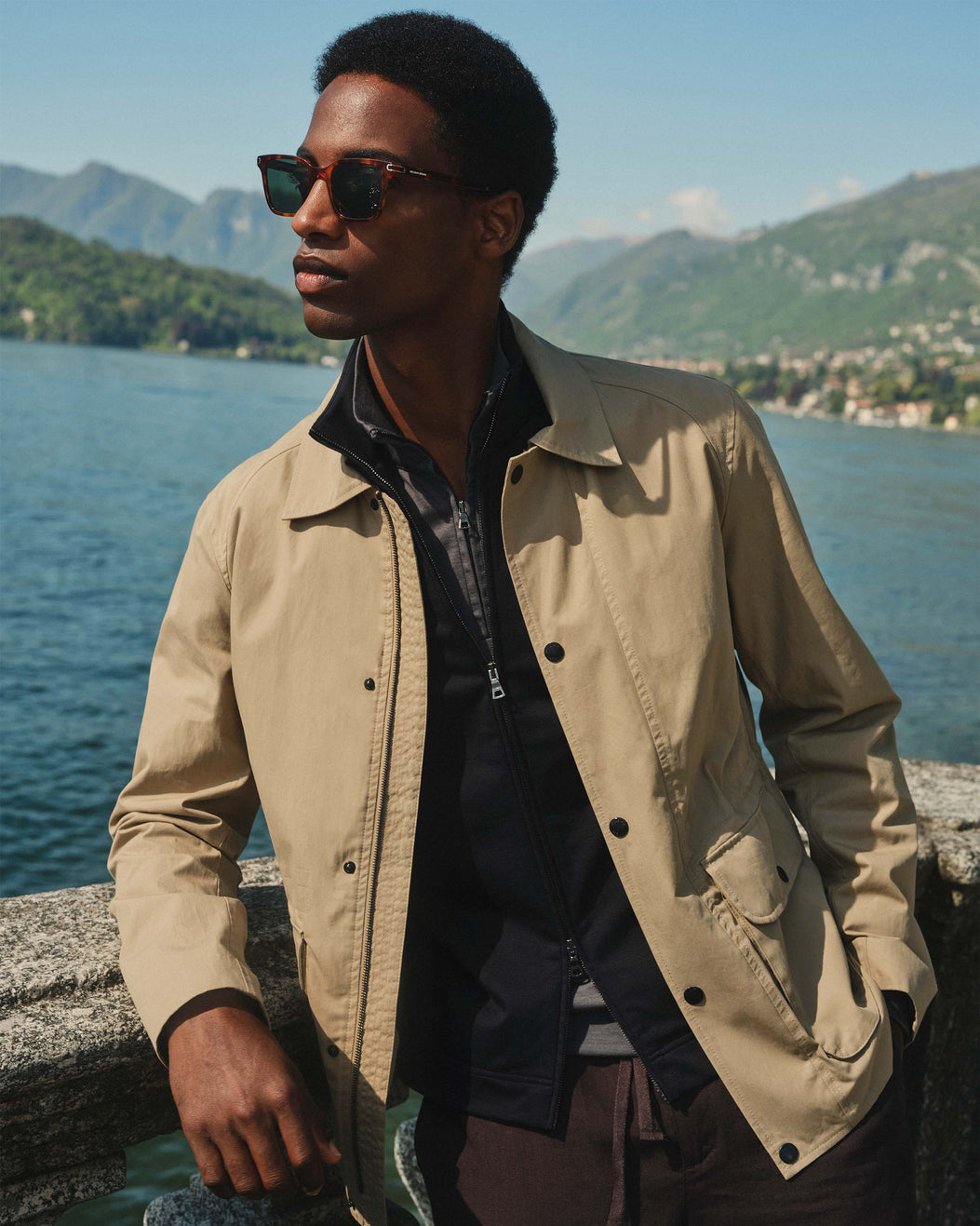 Man wearing a beige jacket and sunglasses by a lake with mountains in the background
