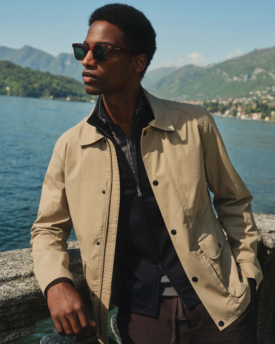 Man wearing a beige jacket and sunglasses by a lake with mountains in the background