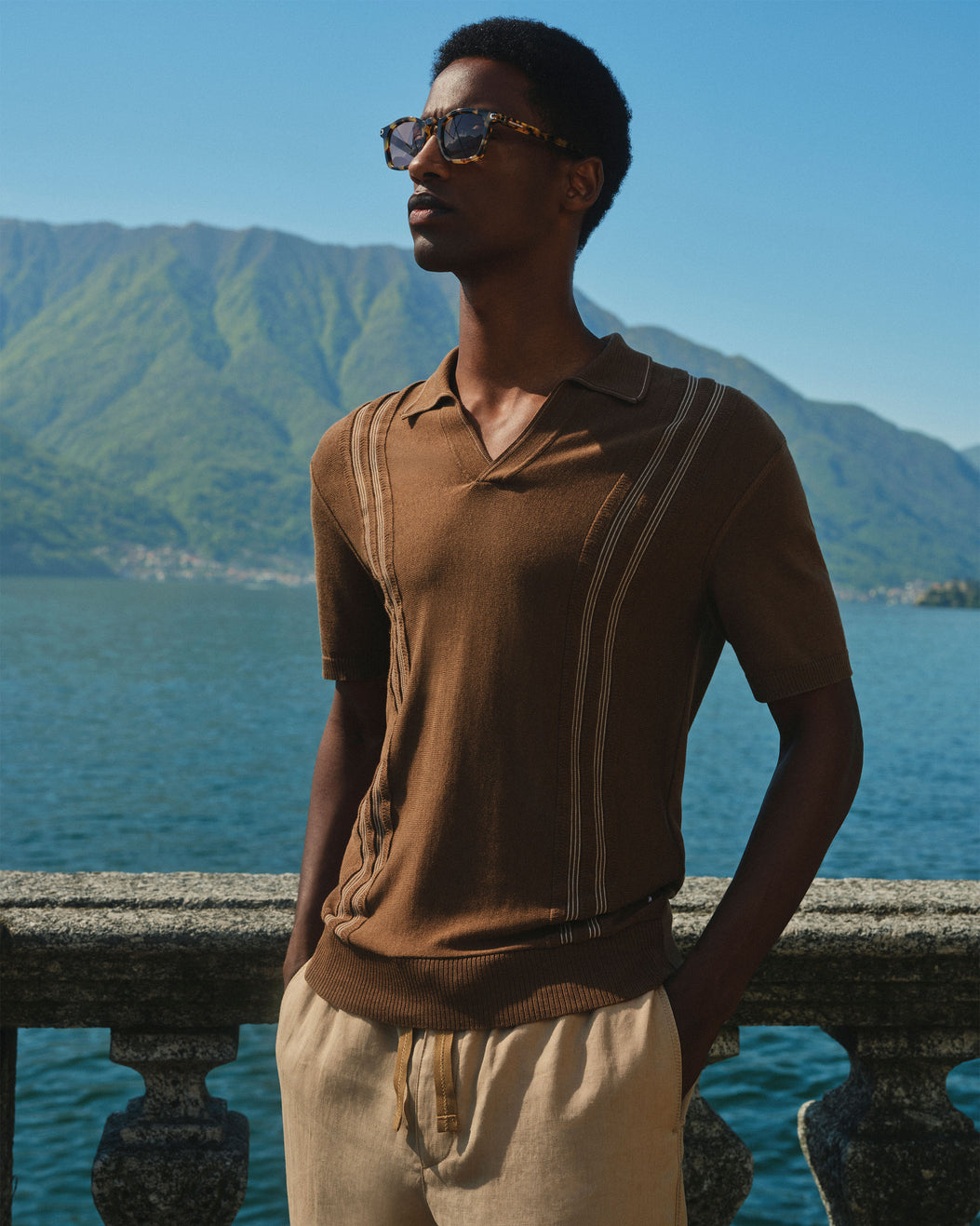 A man wearing a brown short-sleeve knit polo shirt with vertical stripe details and beige drawstring trousers, standing outdoors by a stone railing with a lake and green mountains in the background.