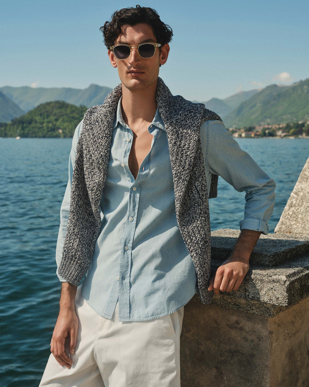 Man wearing sunglasses and a light blue shirt with a gray vest by a lake with mountains in the background
