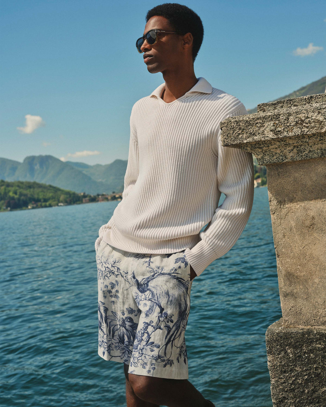 Person wearing a light sweater and patterned shorts by a body of water with mountains in the background