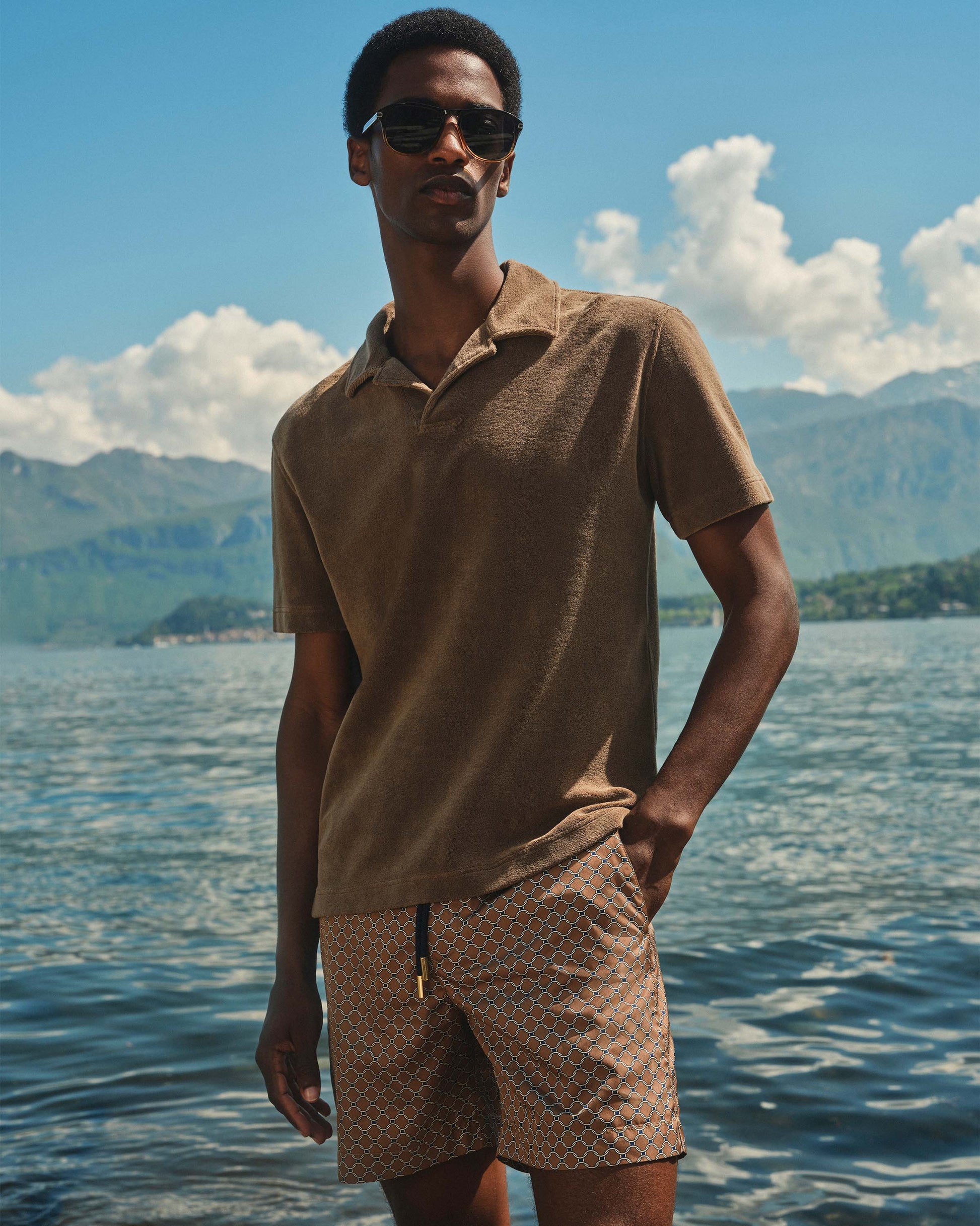 Man wearing sunglasses and a brown shirt and shorts standing by a lake with mountains in the background