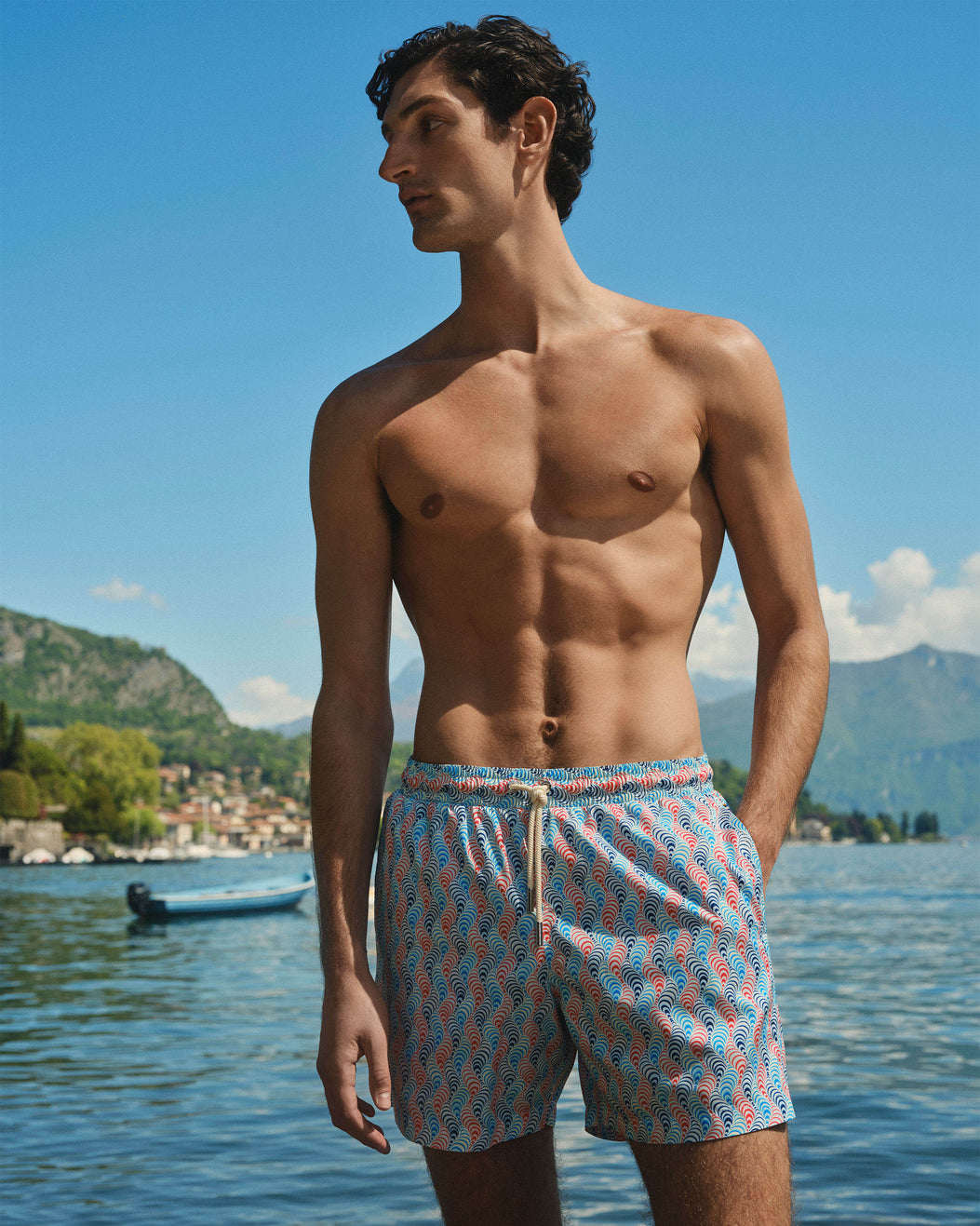 A shirtless man wearing patterned swim trunks stands by a lake with mountains and a boat in the background. The swim trunks have a light blue base with a repeating coral-pink and white abstract pattern.