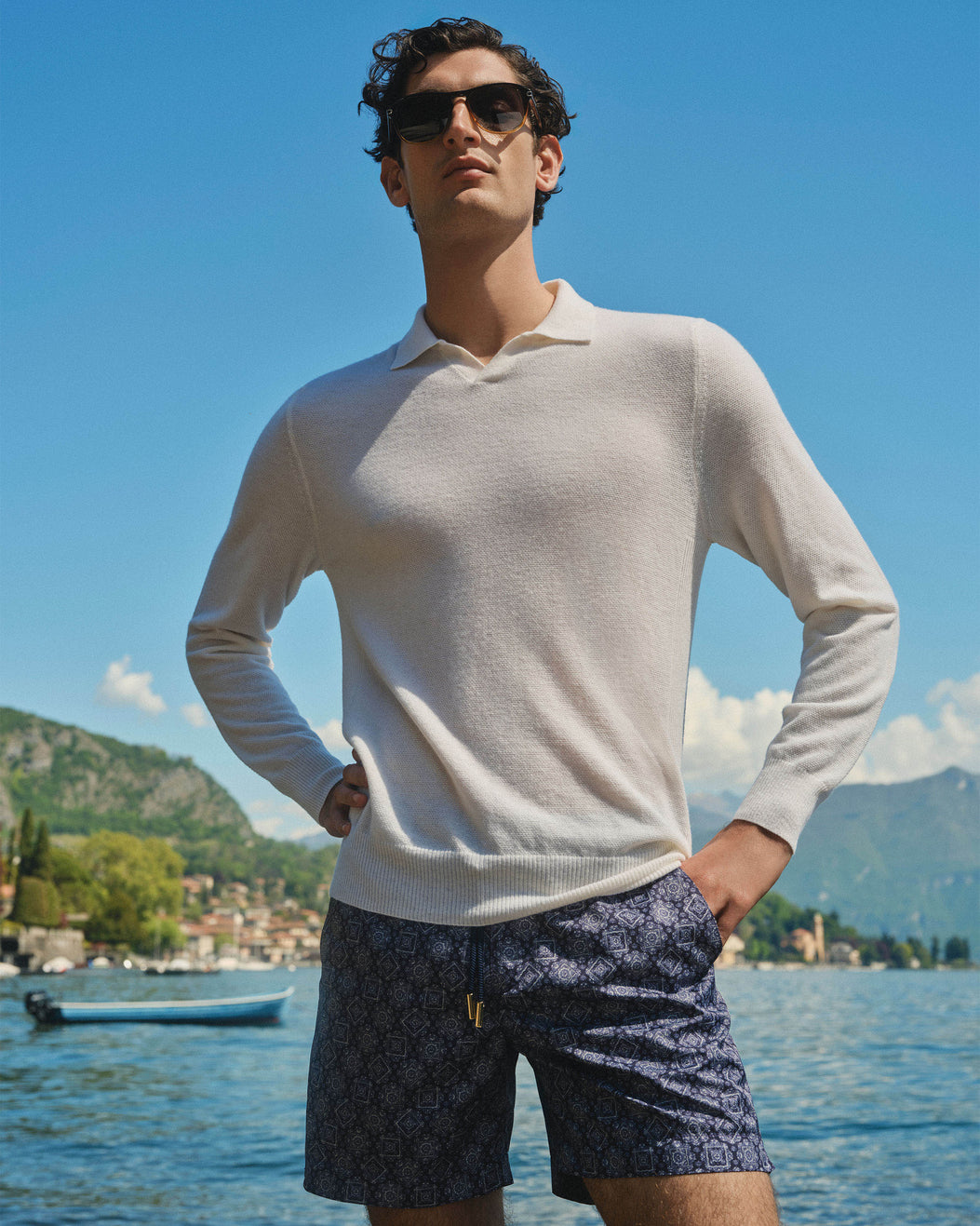 A man wearing a light beige long-sleeve polo sweater and dark patterned shorts stands by a lake with mountains and a boat in the background.