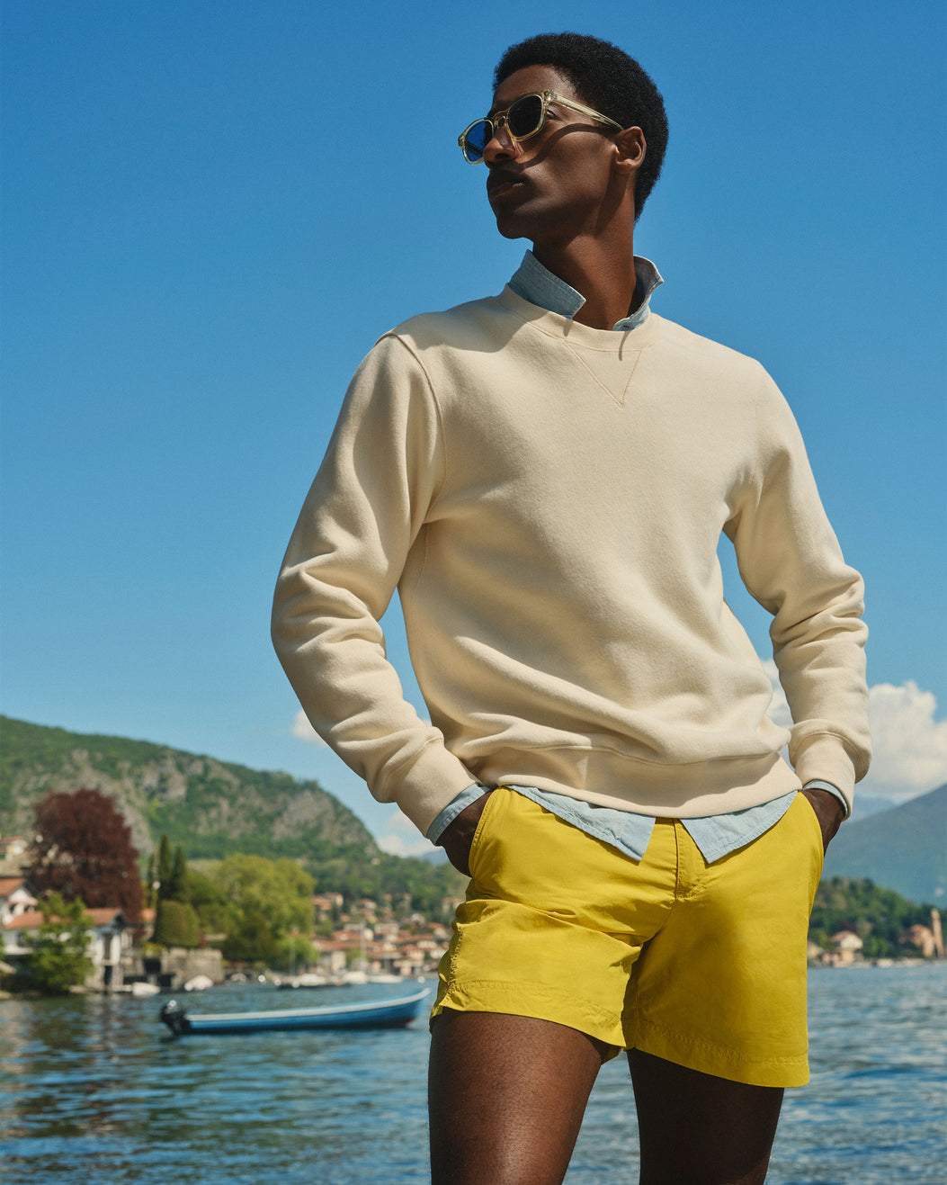 A man wearing a cream quarter-zip sweater over a light blue collared shirt and bright yellow shorts, standing outdoors by a lake with mountains and a boat in the background.